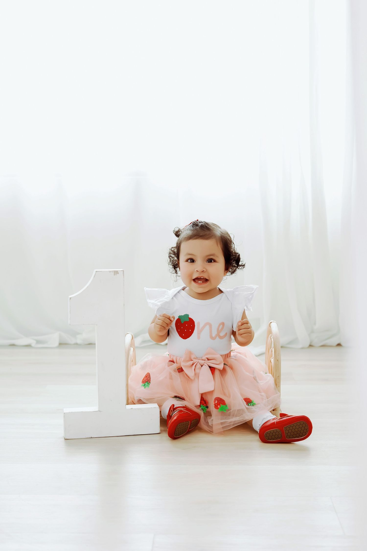 A baby in pink outfit sits on white floor with strawberry decorations in a bright studio setting.