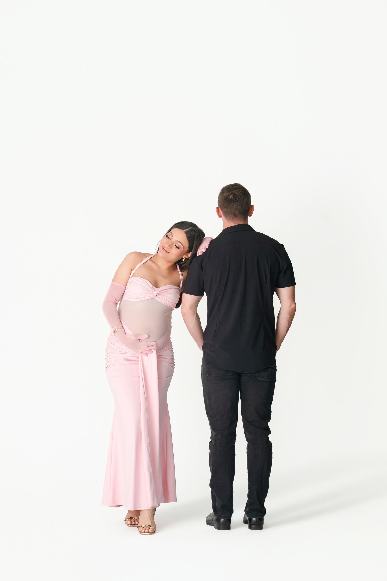 Pregnant woman in pink dress leans on man in black outfit during maternity photo shoot on white background.