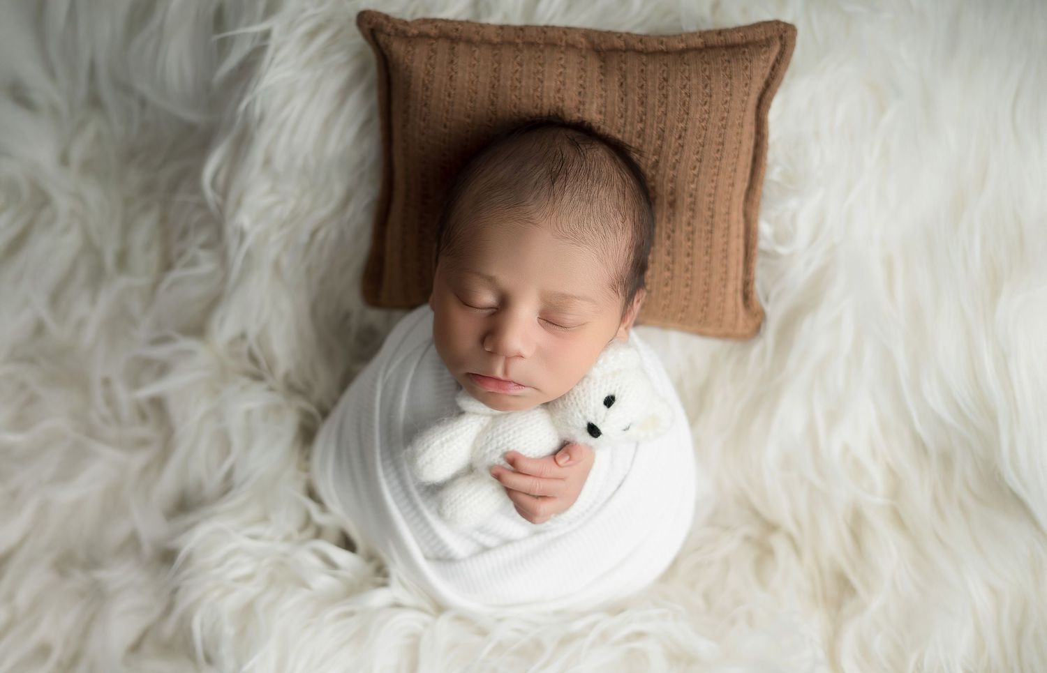 Newborn baby swaddled in white wrap sleeping peacefully on fluffy rug holding a white teddy bear on brown pillow.