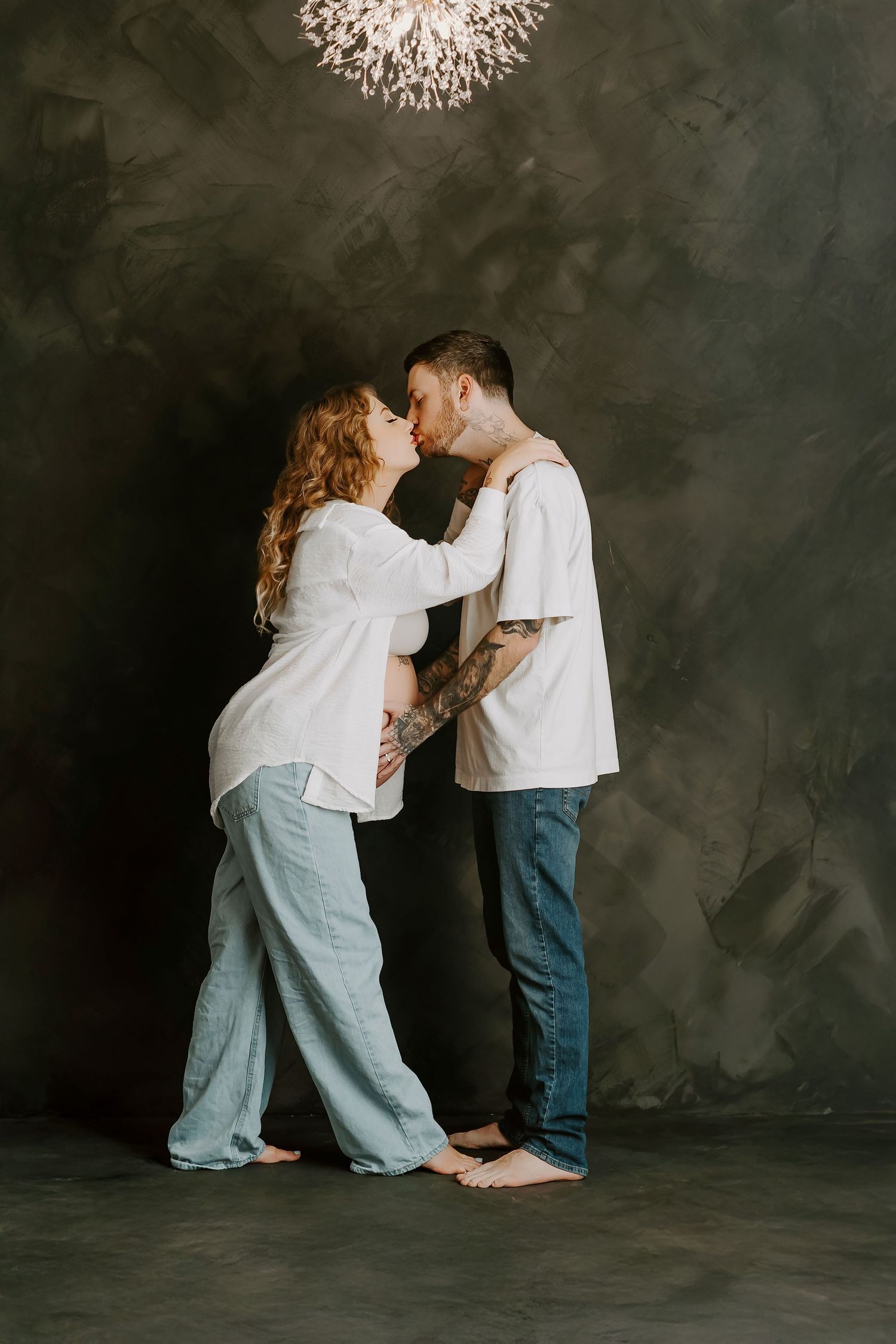 Pregnant couple sharing a tender kiss in a studio, wearing casual white tops and jeans, with tattoos visible.