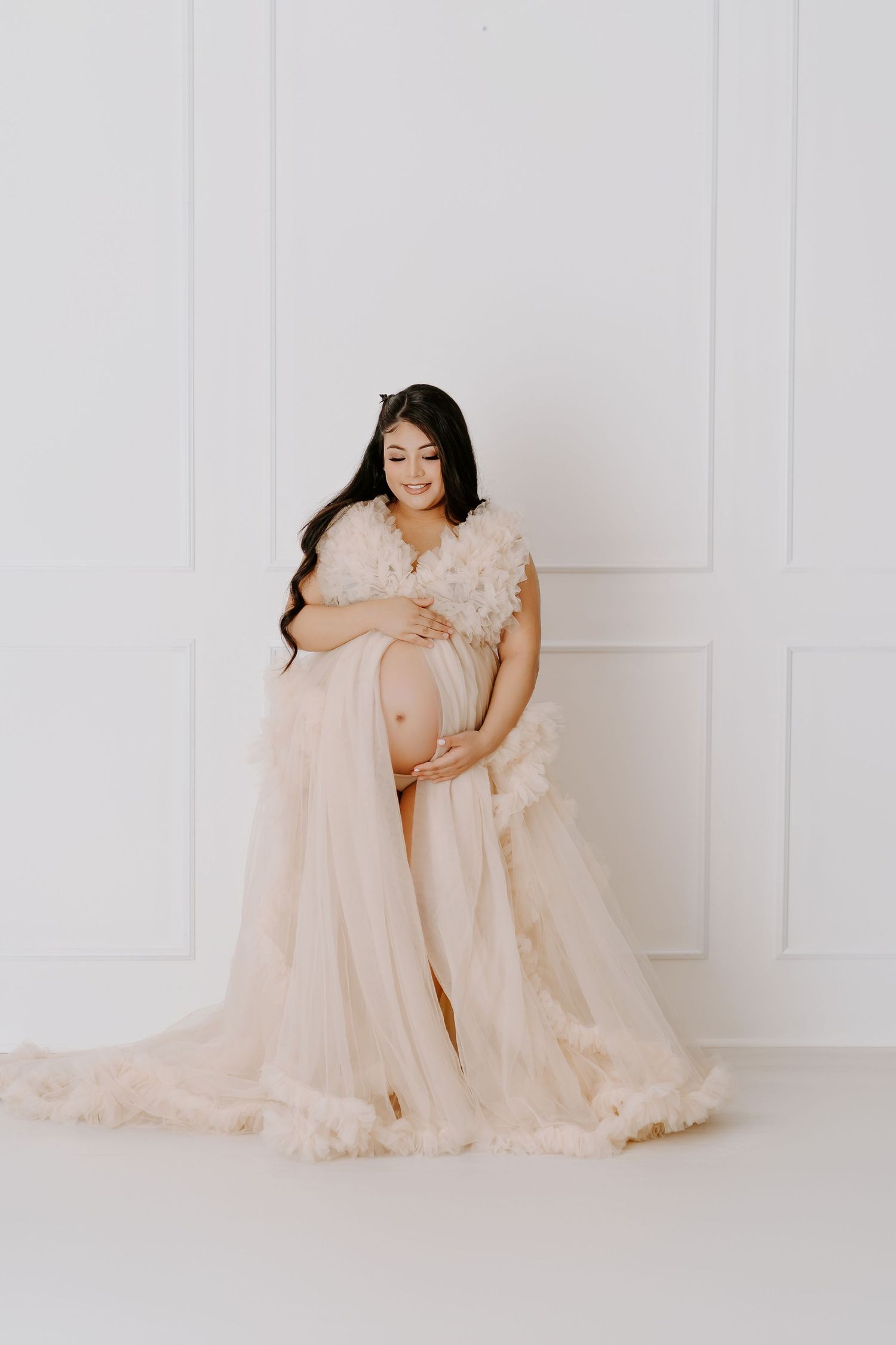 Pregnant woman in elegant blush pink tulle maternity gown cradling baby bump against white studio backdrop.