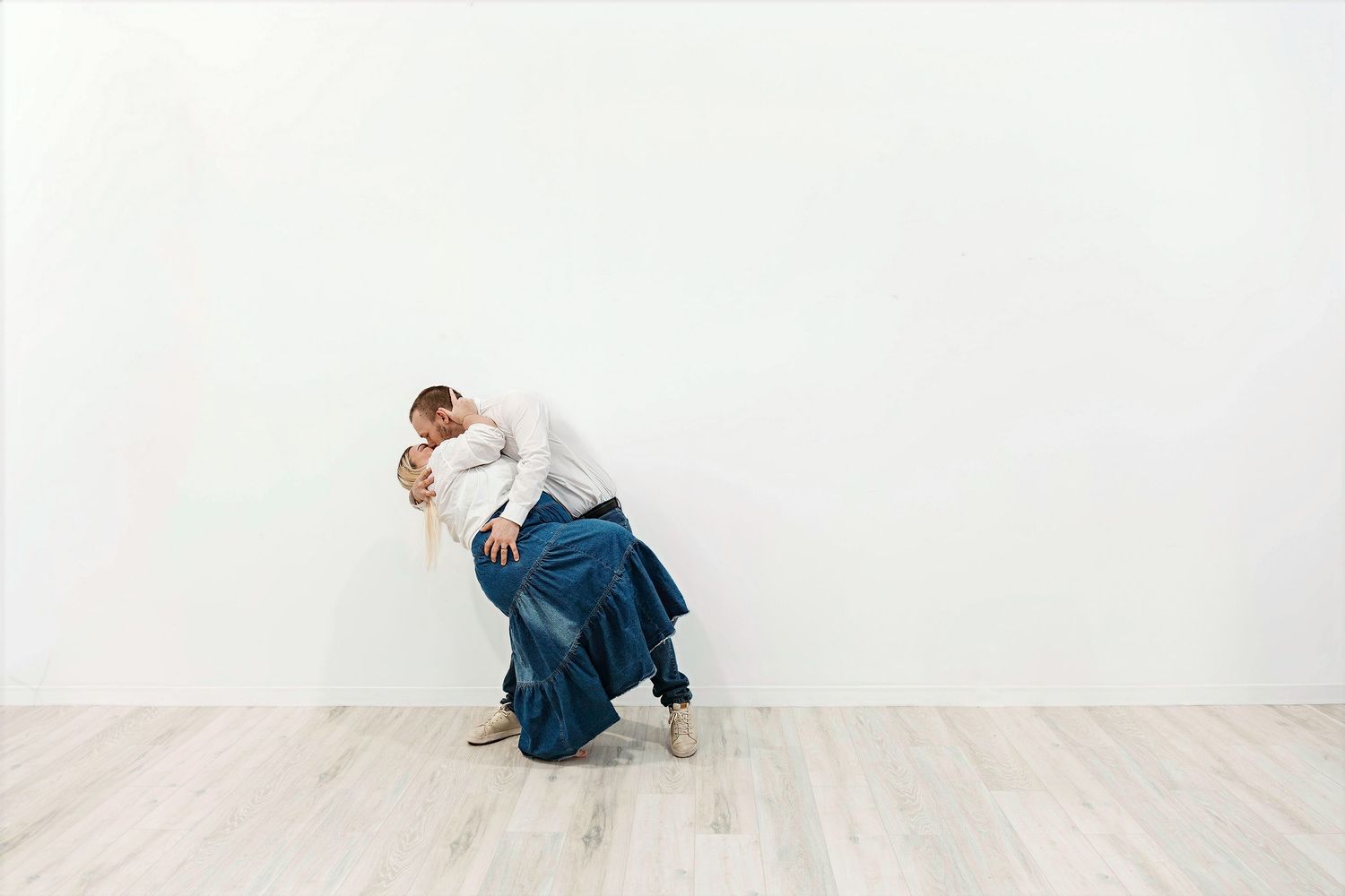 Person in blue denim crouches and curls into a ball position against white wall in minimalist setting.