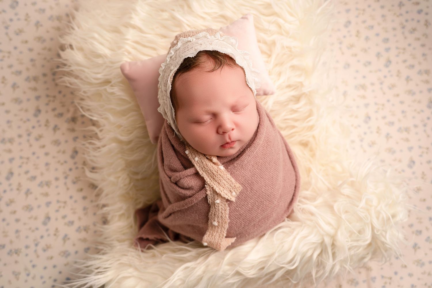Newborn baby swaddled in mauve wrap with lace bonnet, sleeping on fluffy cream fur blanket with pink pillow.