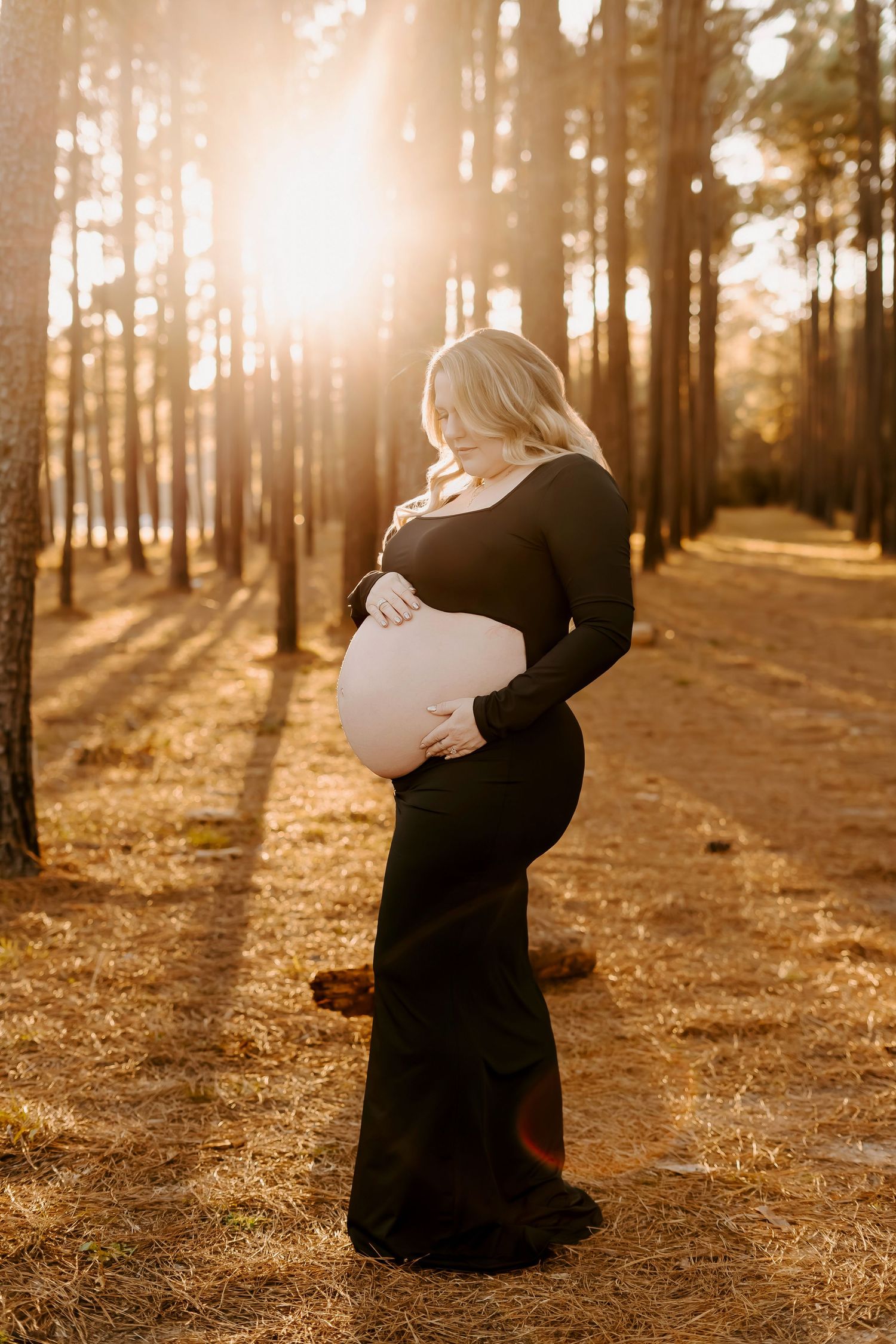 A pregnant woman in a black dress stands in a sunlit forest path during golden hour.