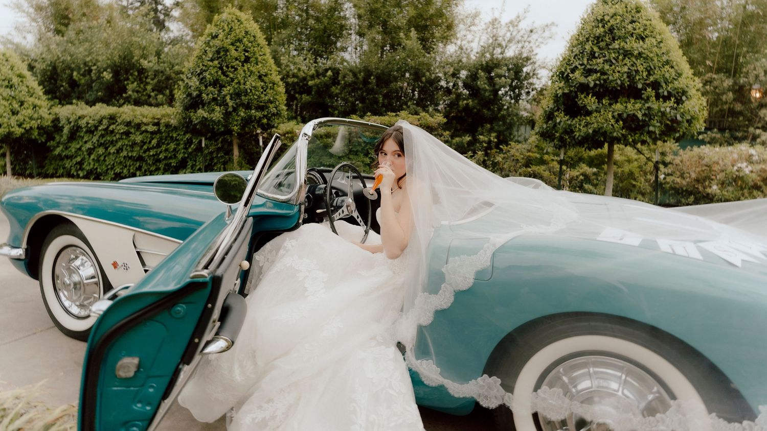Bride in white wedding gown and veil seated in a vintage teal convertible car surrounded by lush greenery.