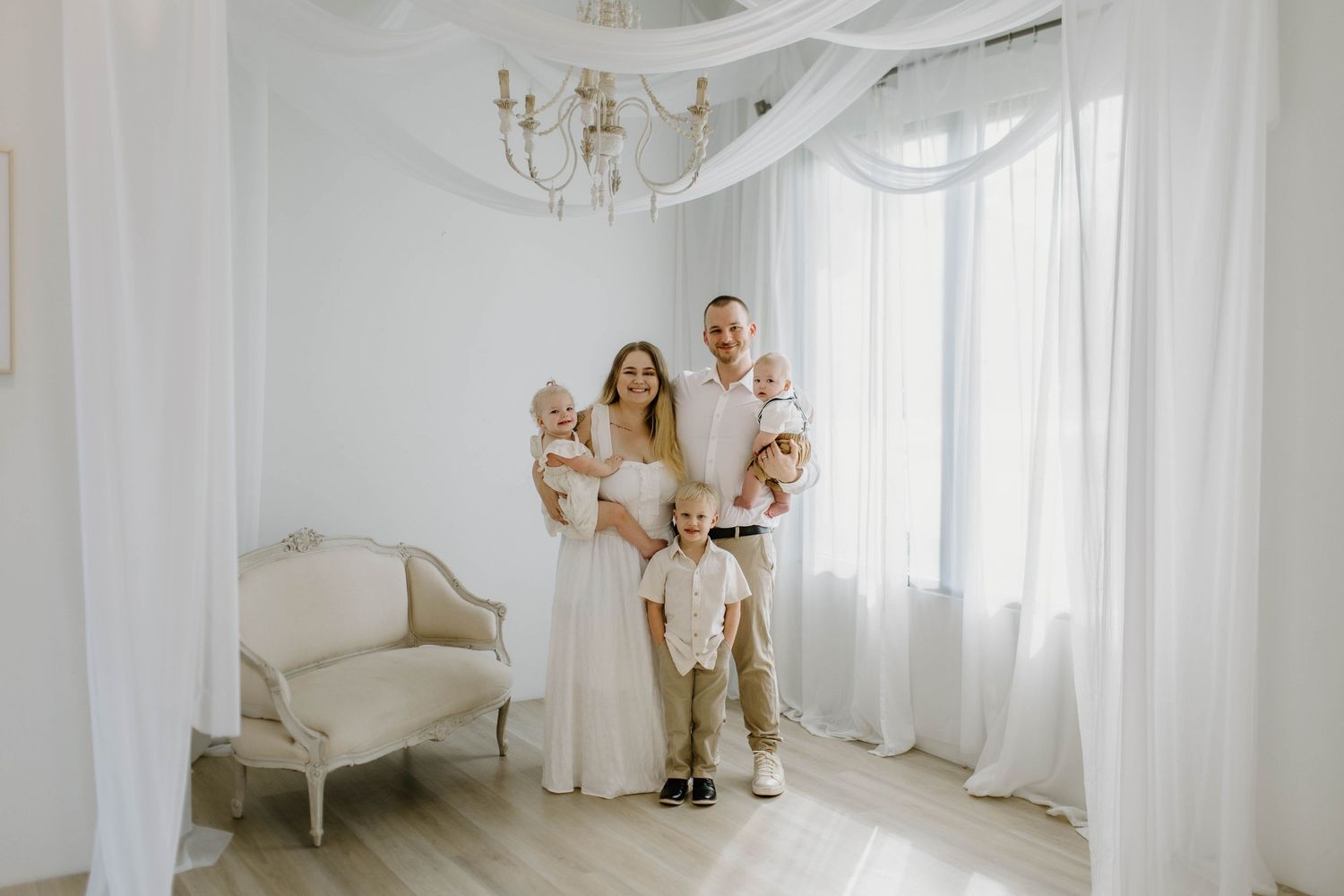 A family in white outfits poses together in a bright minimalist room with sheer curtains and an elegant chandelier.