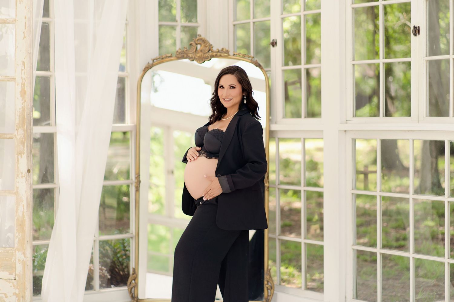 Pregnant woman in black blazer and pants poses elegantly before ornate gold mirror in bright white room.