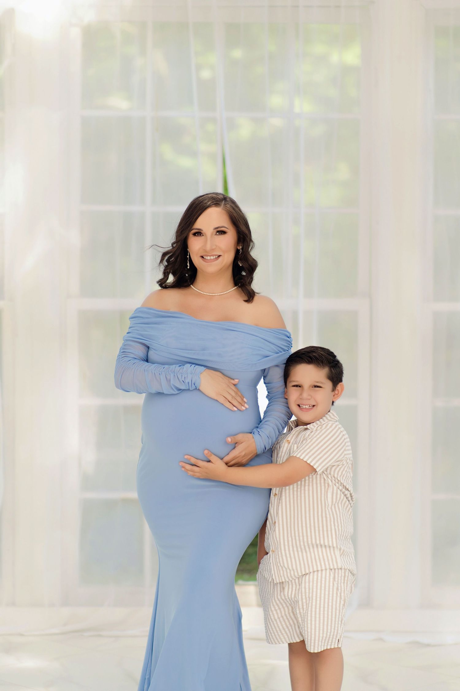 Pregnant woman in blue off-shoulder gown poses with young boy in striped outfit during maternity photo session.