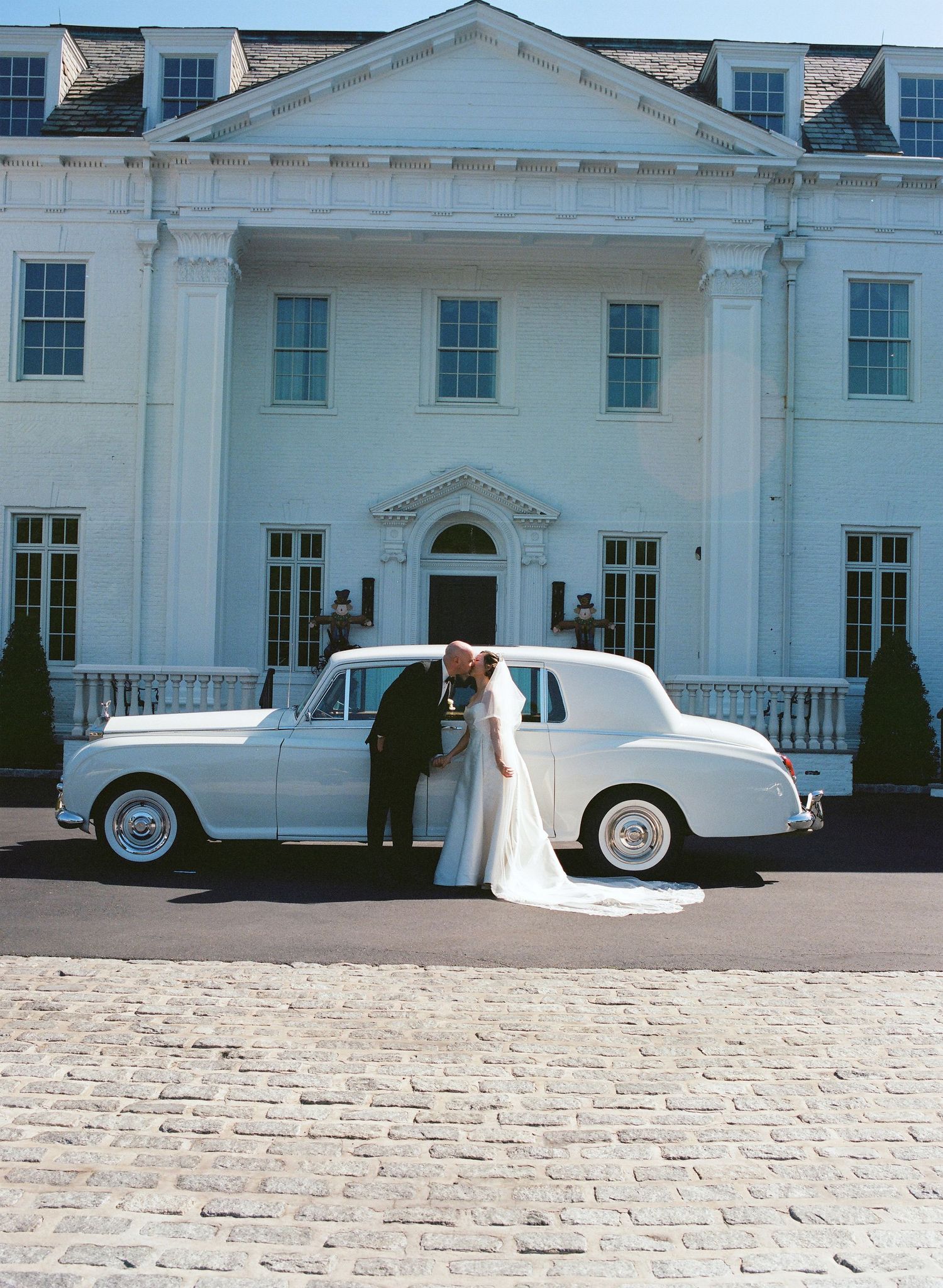 Classic white vintage car parked in front of elegant white mansion.