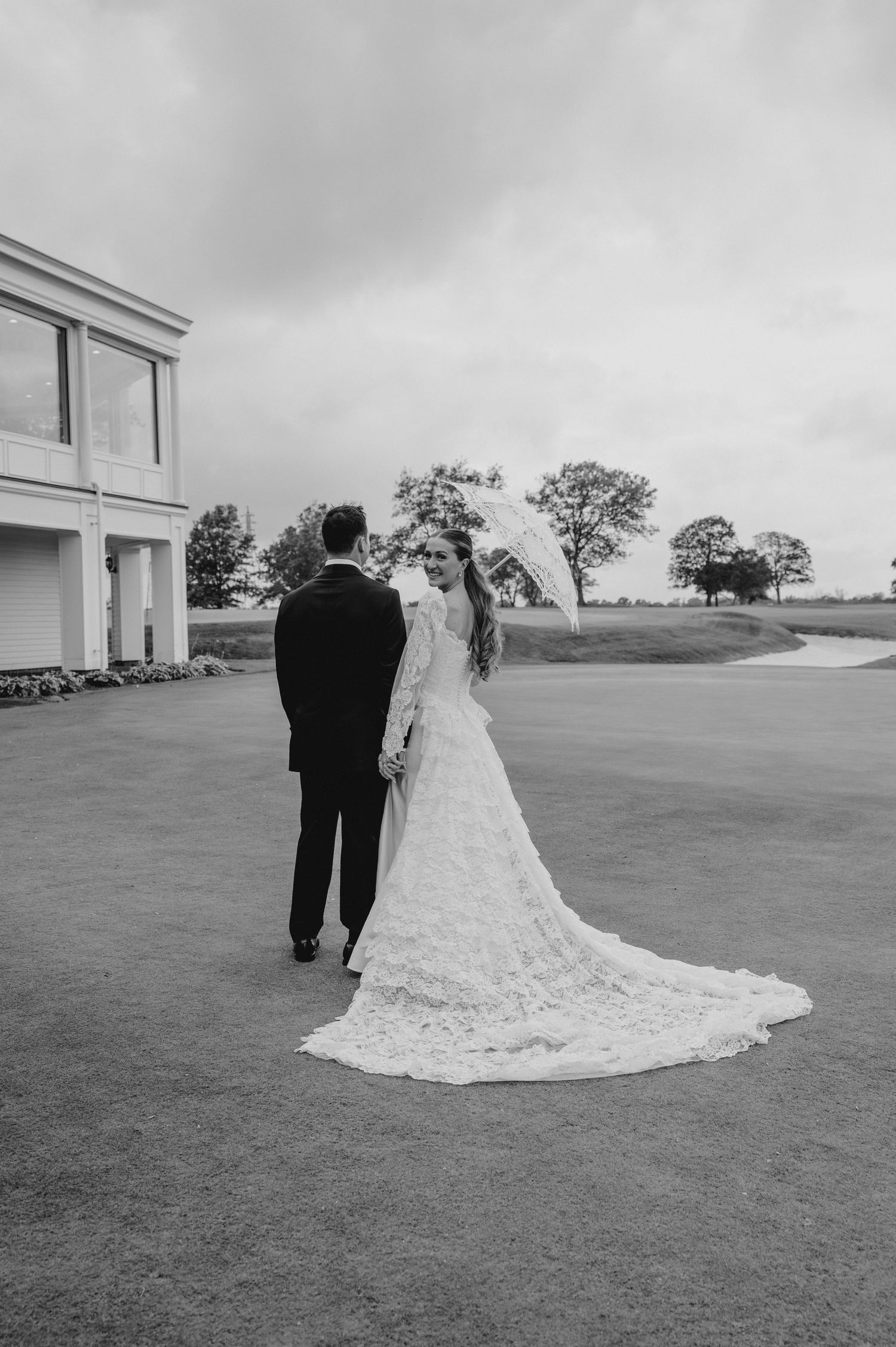 Wedding couple walking across expansive lawn with long lace train trailing behind.