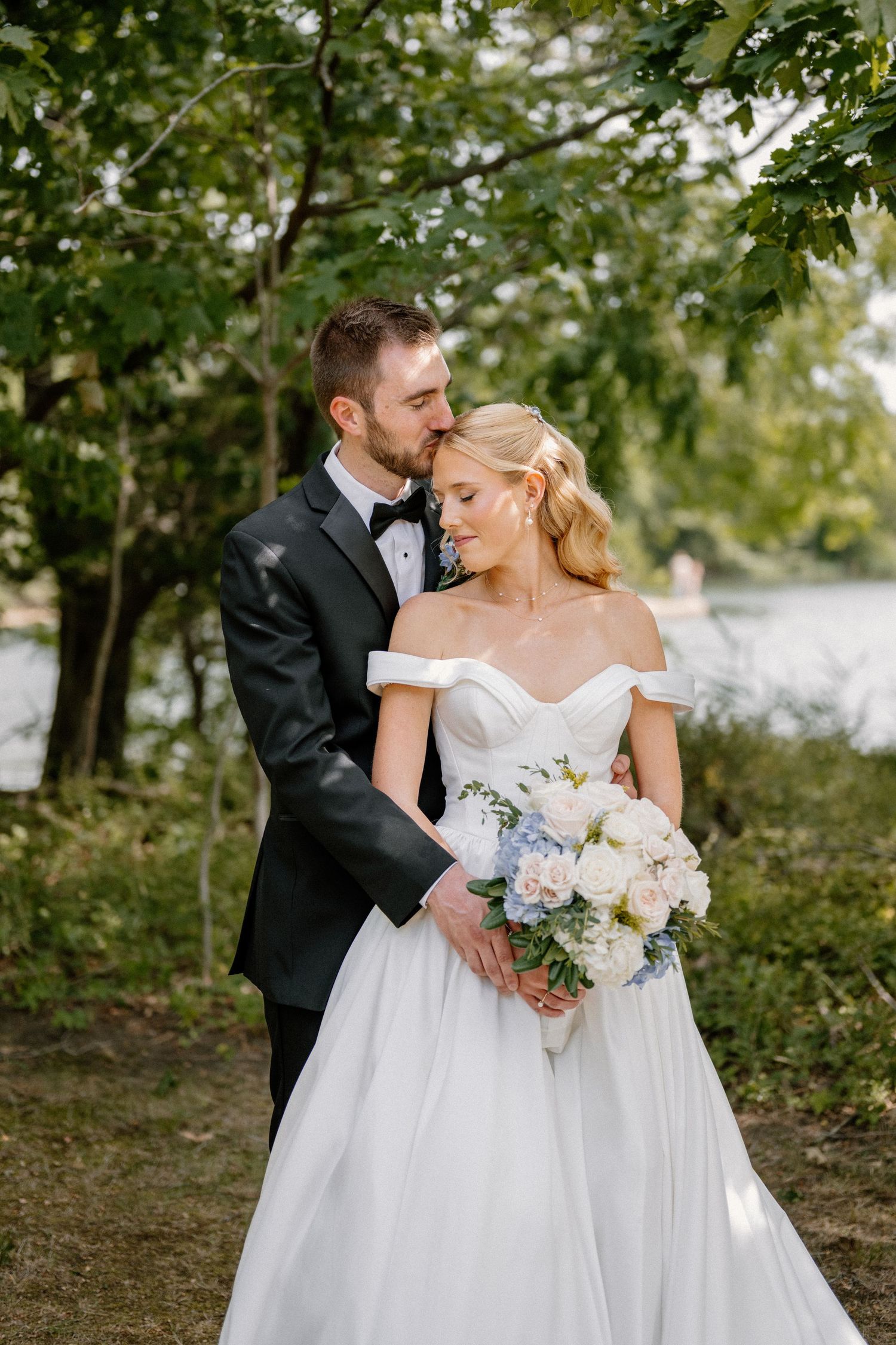 Groom in black tuxedo kissing bride in off-shoulder white gown holding pink bouquet outdoors near trees.