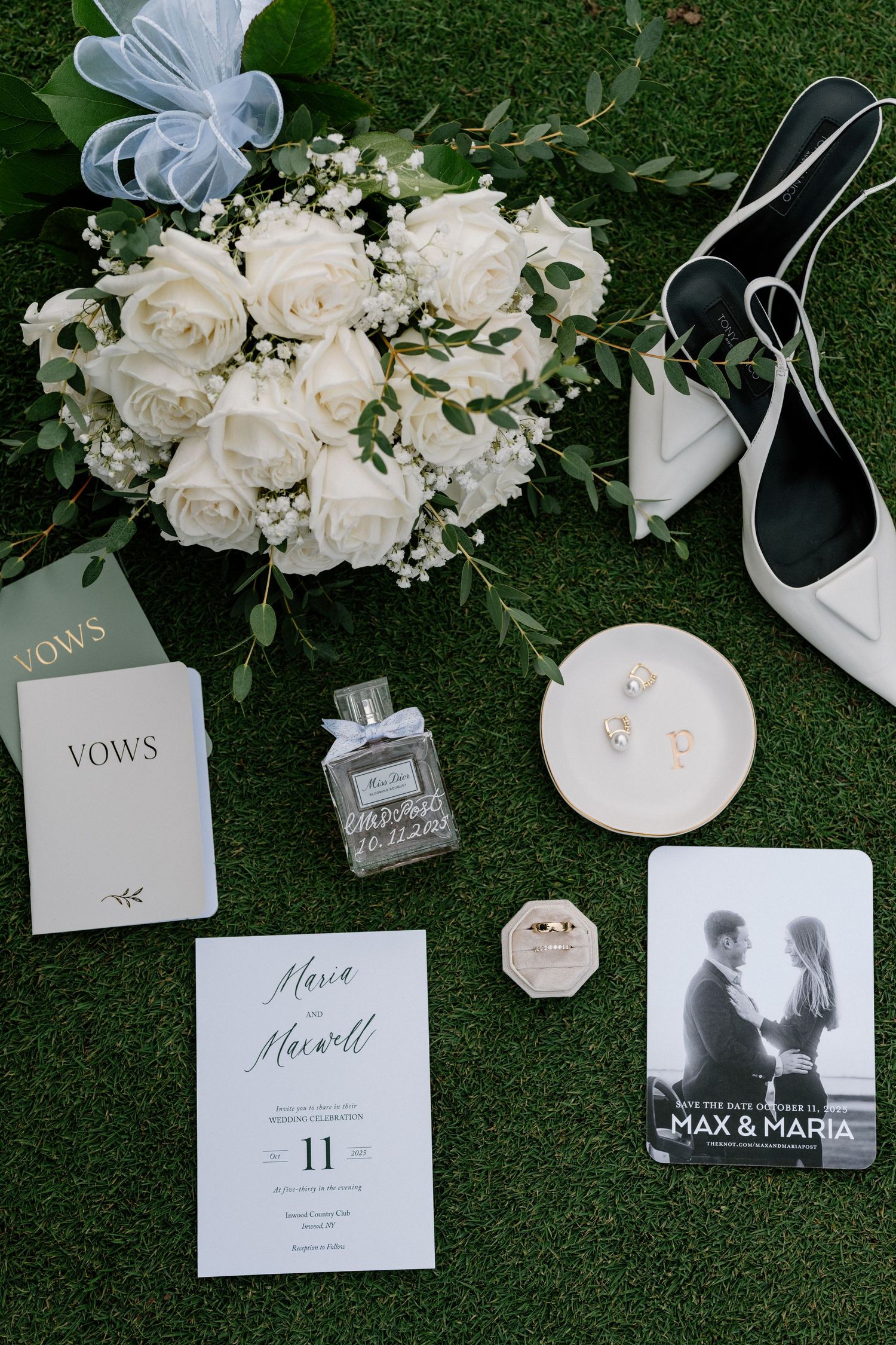 Wedding details flatlay with white roses bouquet, vows book, perfume, invitation and bridal accessories on grass background.