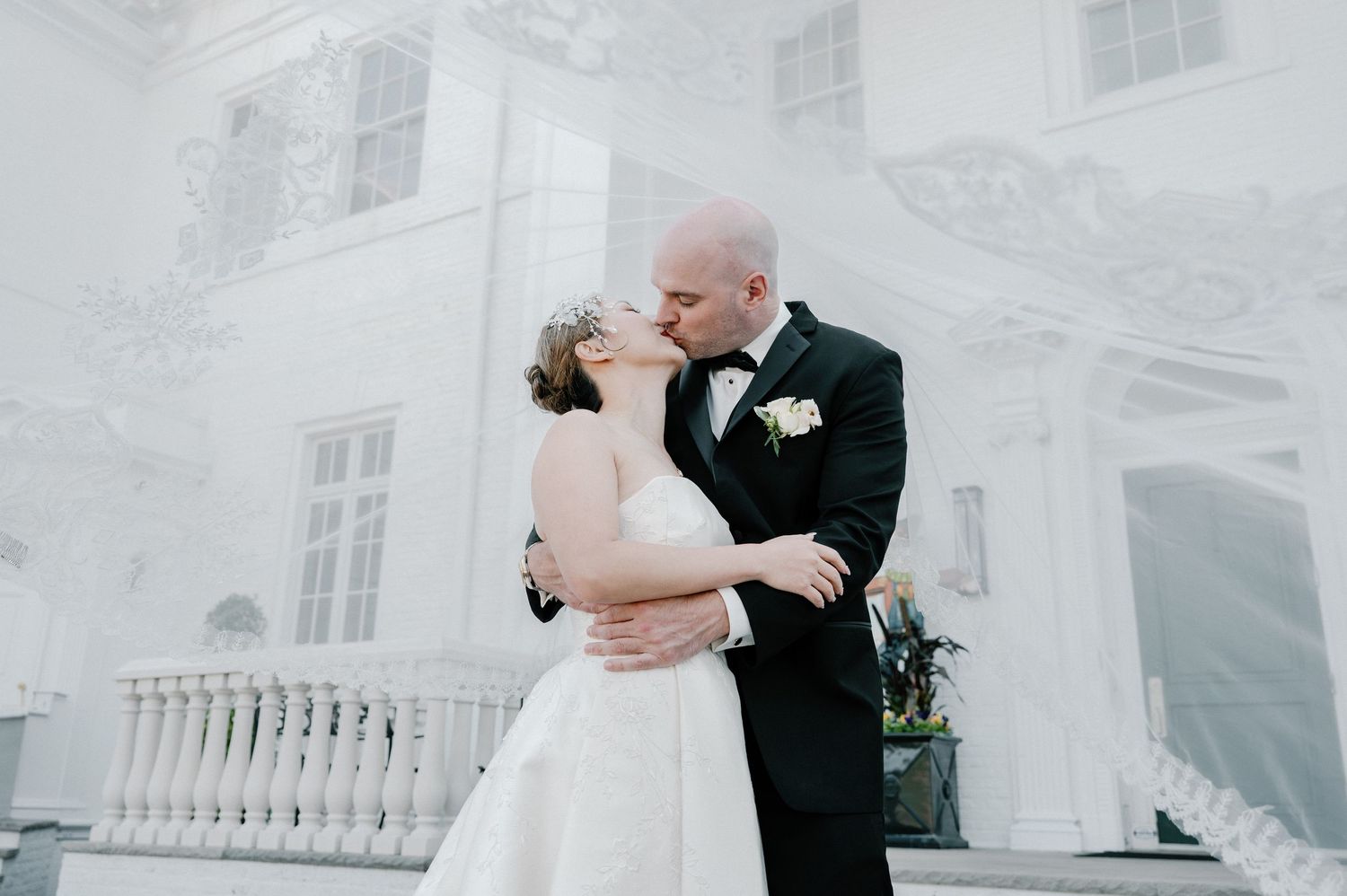 Bride and groom sharing a romantic kiss in front of an elegant white mansion during their wedding day.