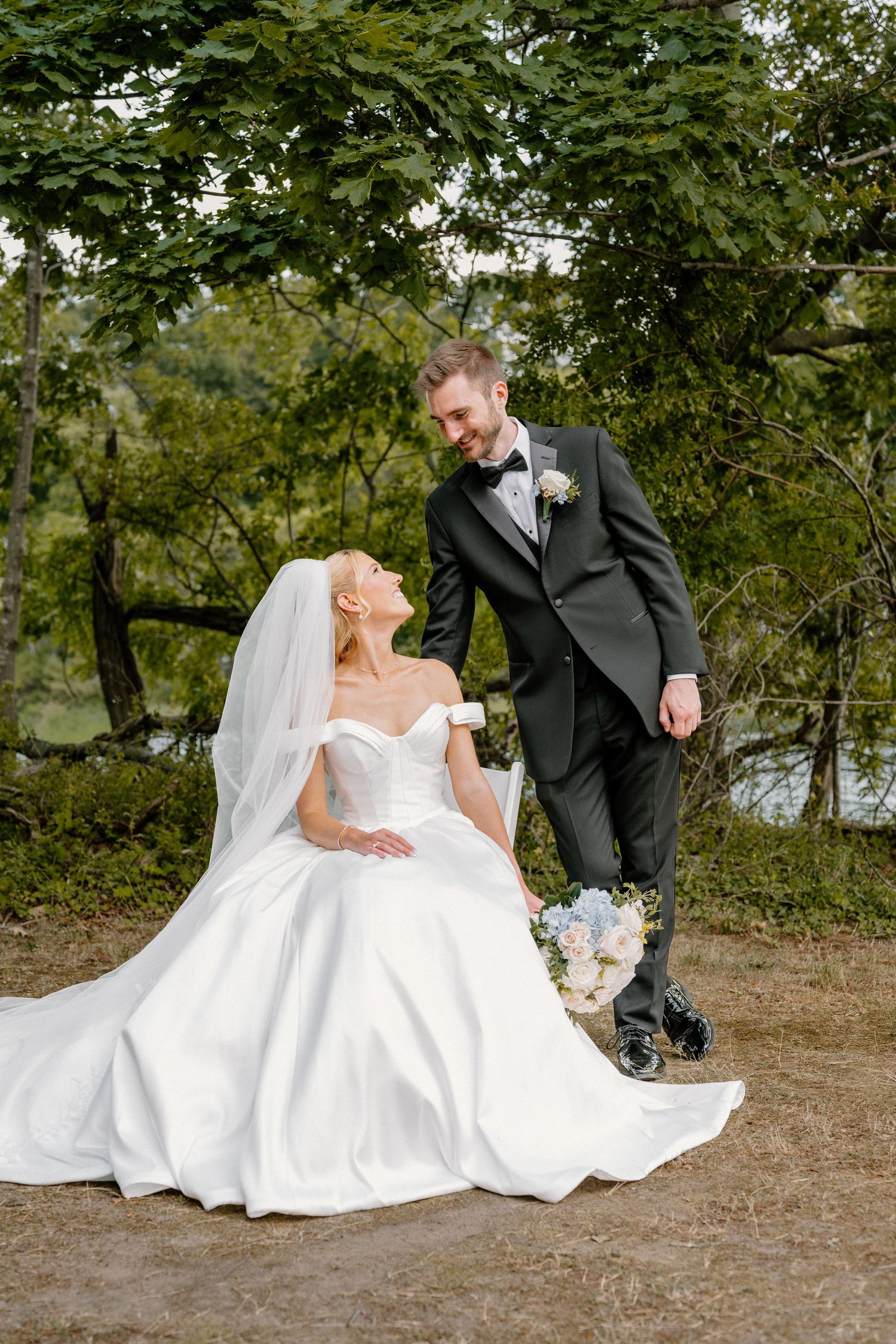A bride in a white gown sits outdoors while her groom in a black suit stands beside her, both smiling at each other.