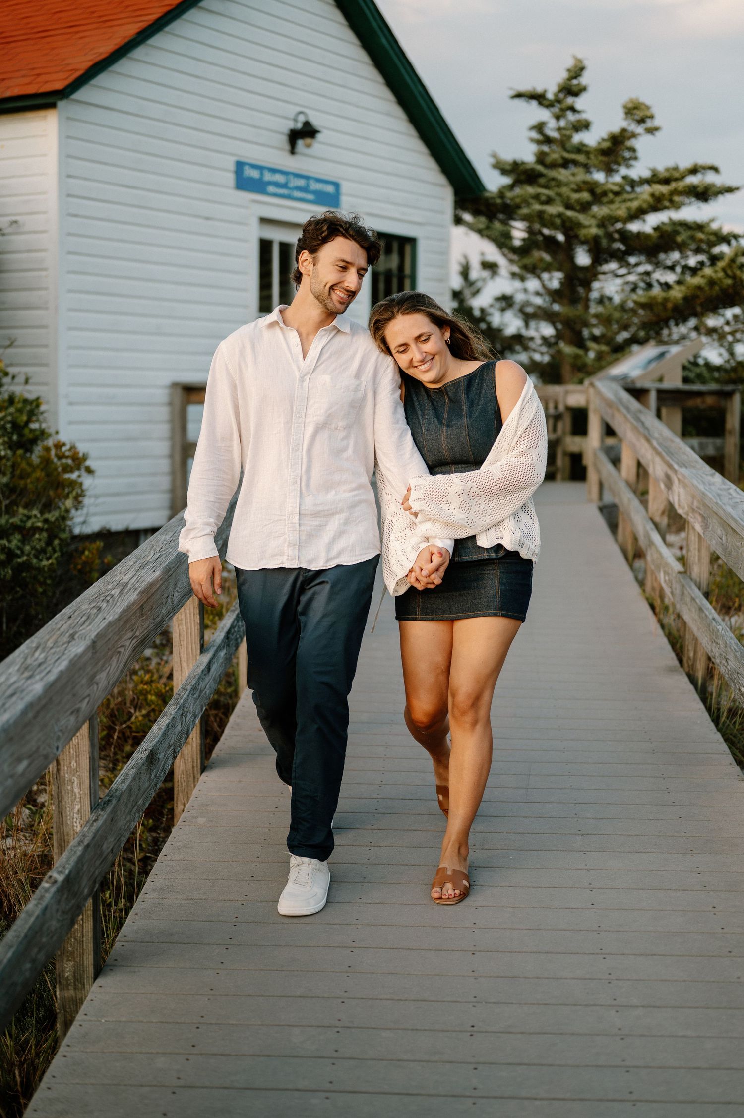 A couple walking together on a wooden boardwalk near a white coastal building.