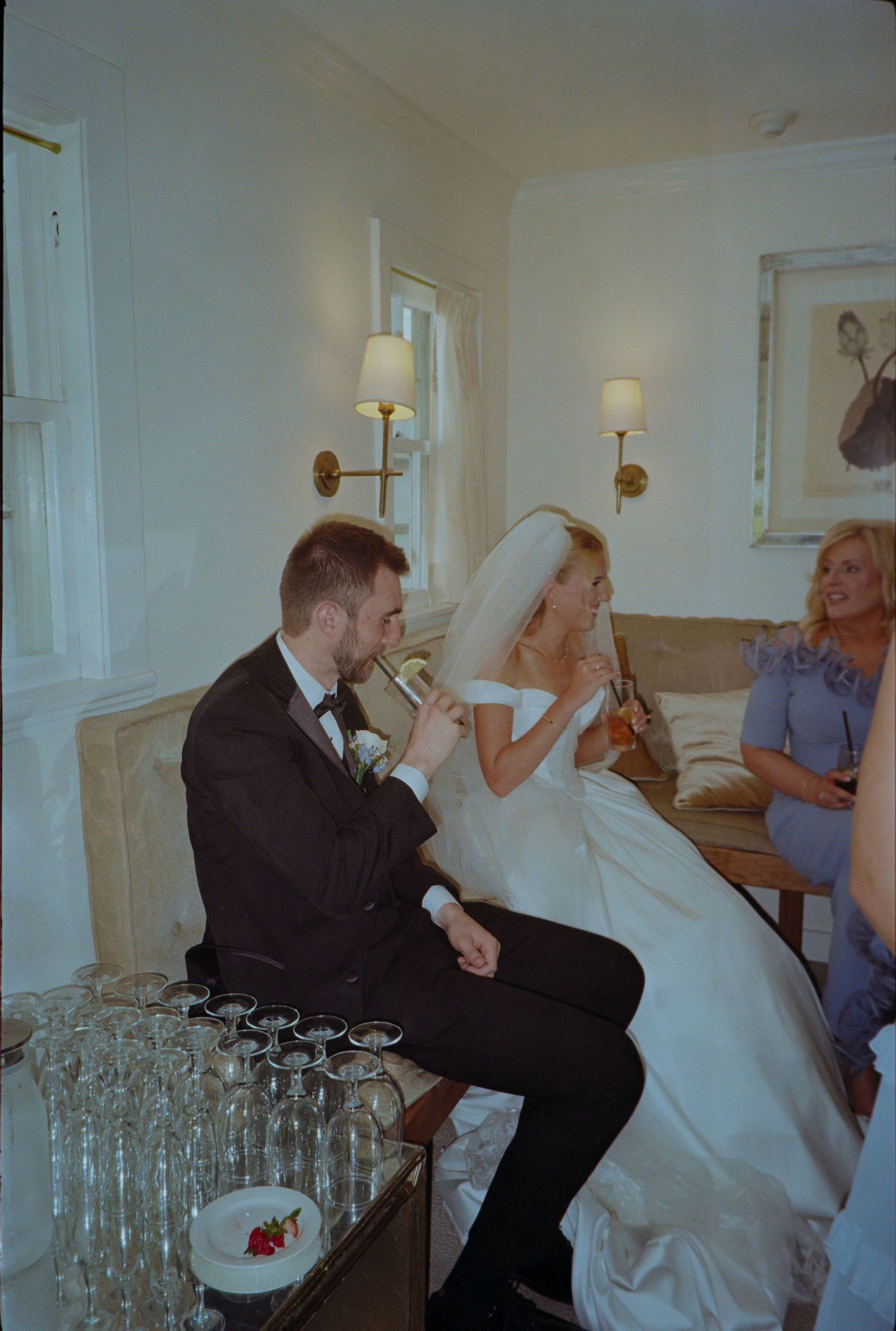 Bride and groom share a champagne toast at their wedding reception, seated together in an elegant white room.