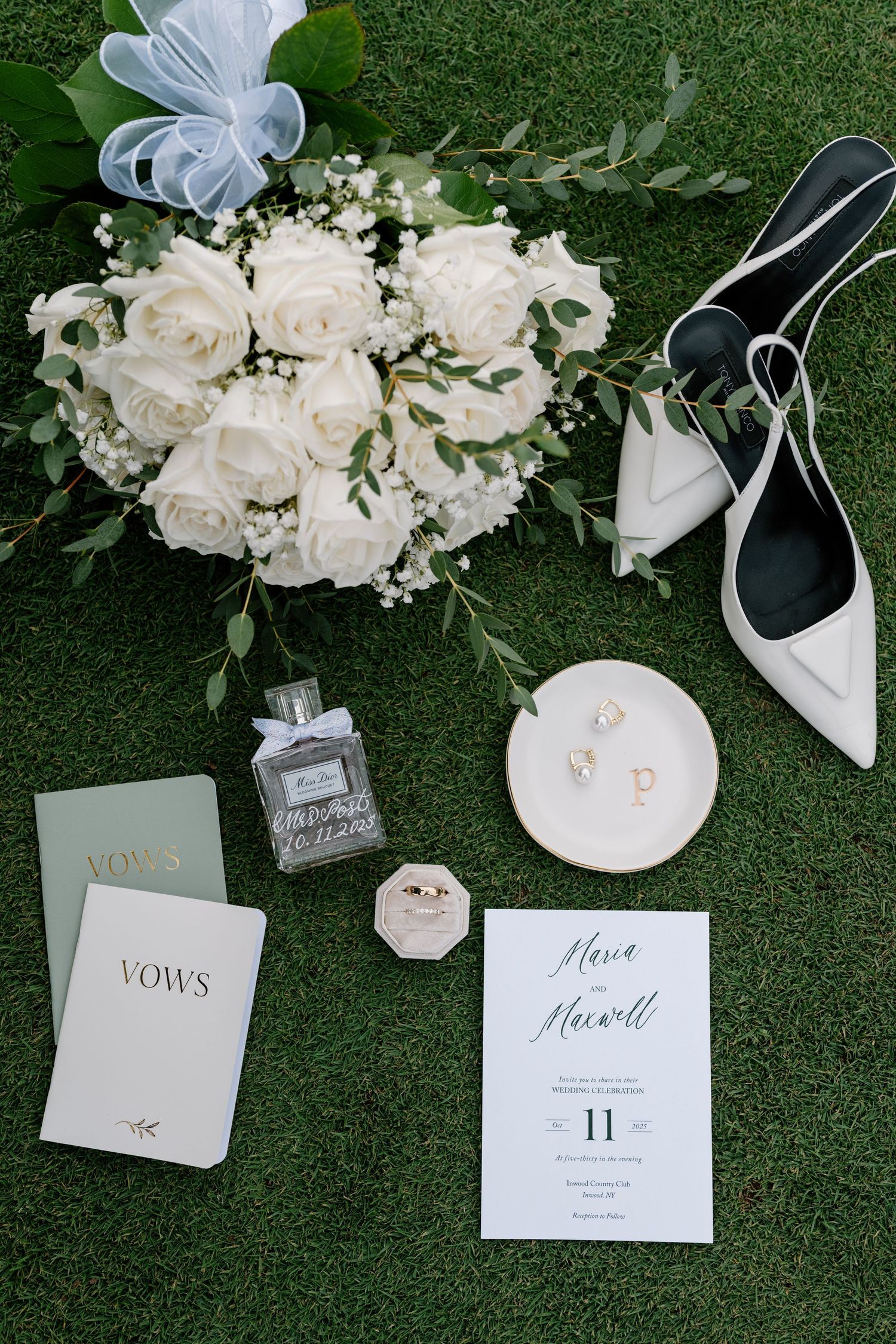 Wedding details flatlay with white roses bouquet, bridal shoes, vows book, perfume and invitation on green grass.