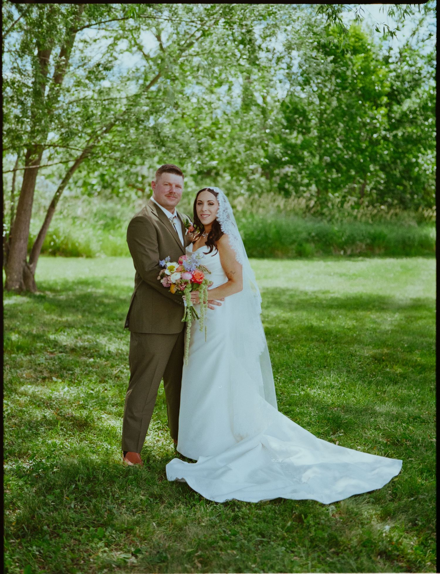Bride and groom posing together outdoors on lush green grass, bride holding colorful bouquet, wearing white gown with long train.
