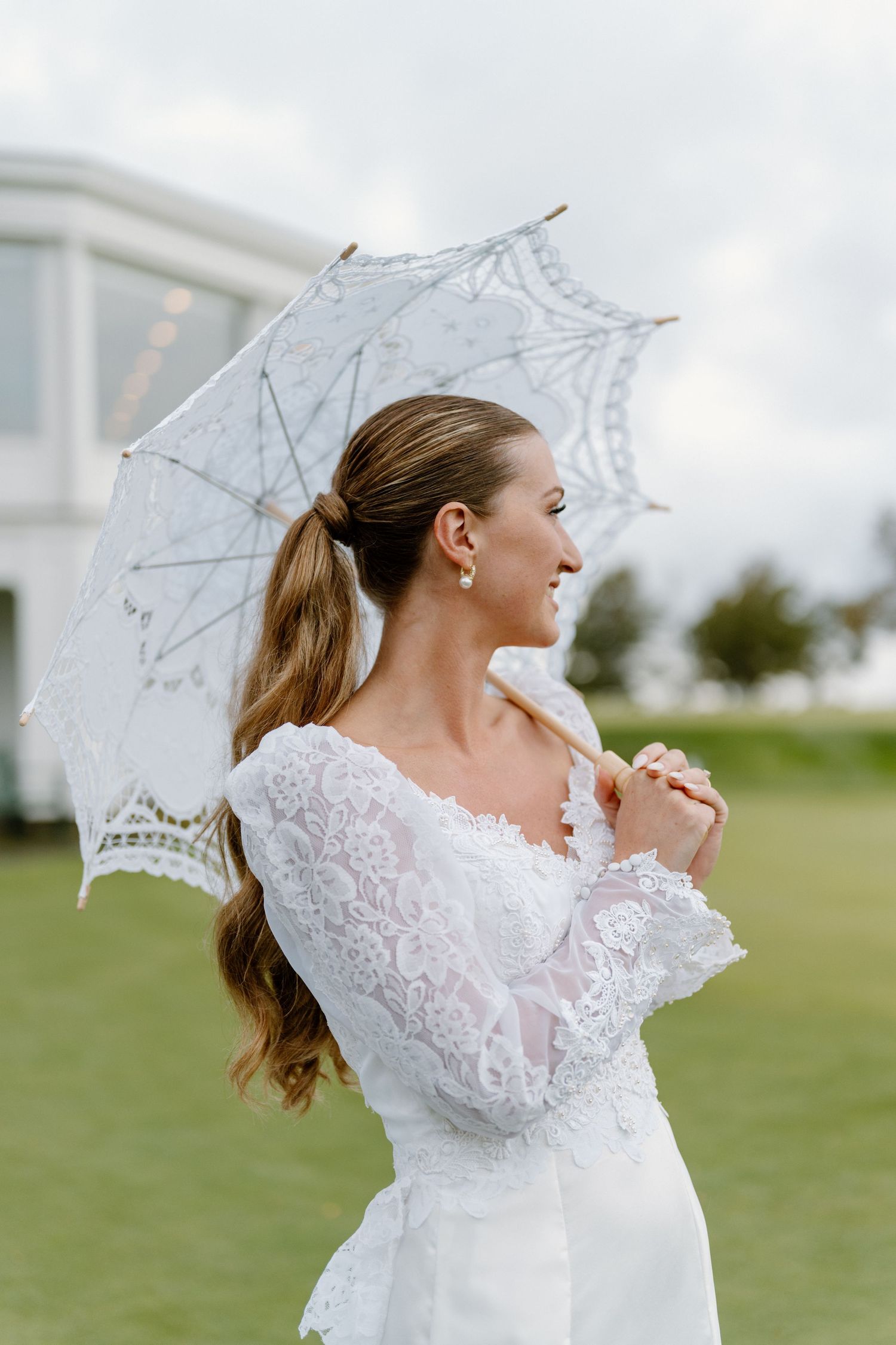 Bride in lace wedding dress holding white lace parasol umbrella outdoors, smiling elegantly on green lawn.
