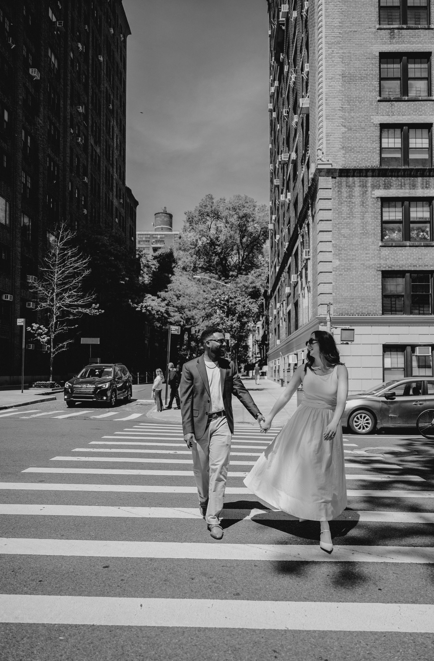 A couple holds hands crossing a zebra crossing in a New York City street, dressed in elegant wedding attire.