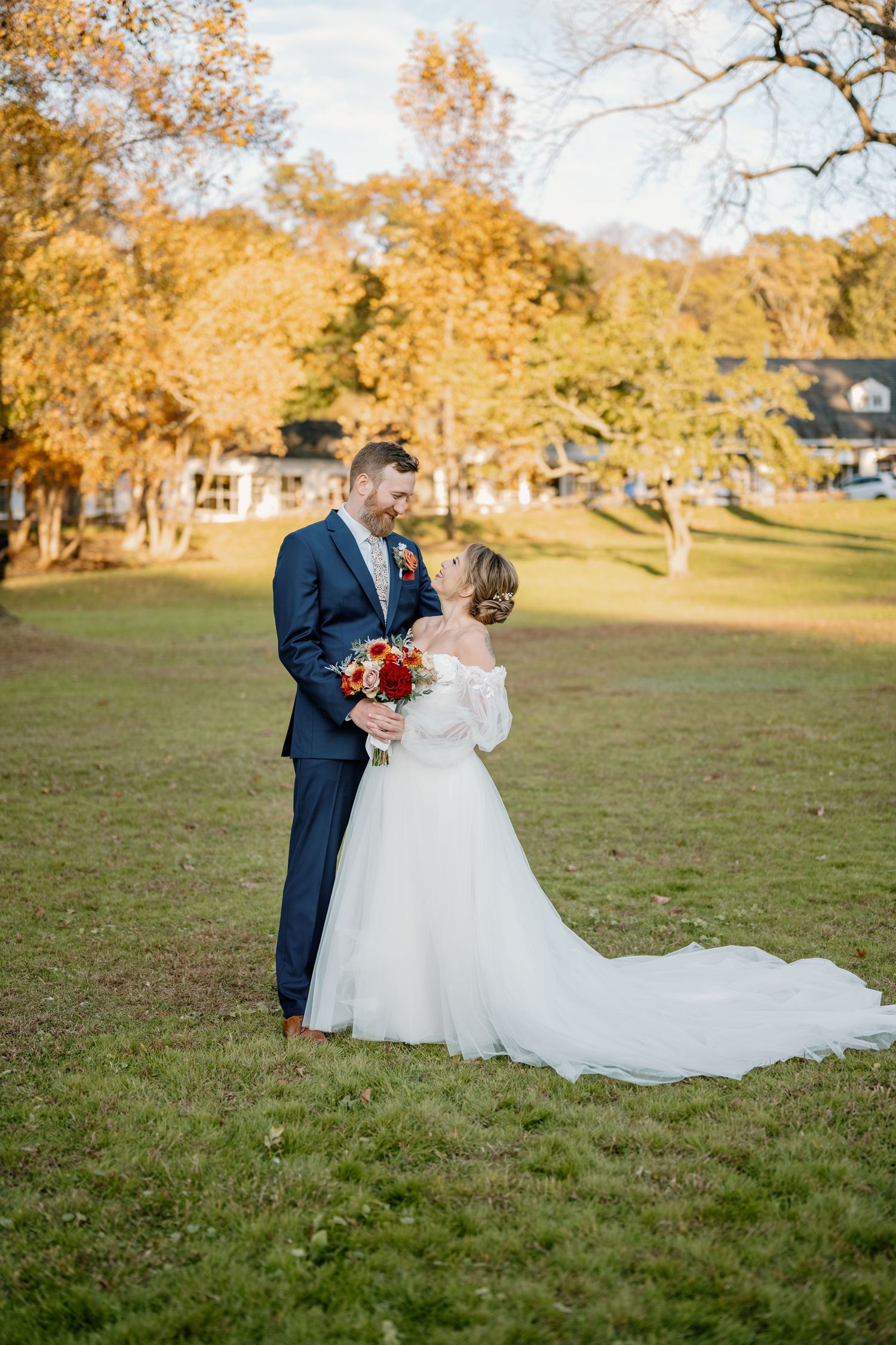 Bride and groom embrace in a sunlit autumn field, she holds a red bouquet in a white gown, he wears a navy suit.