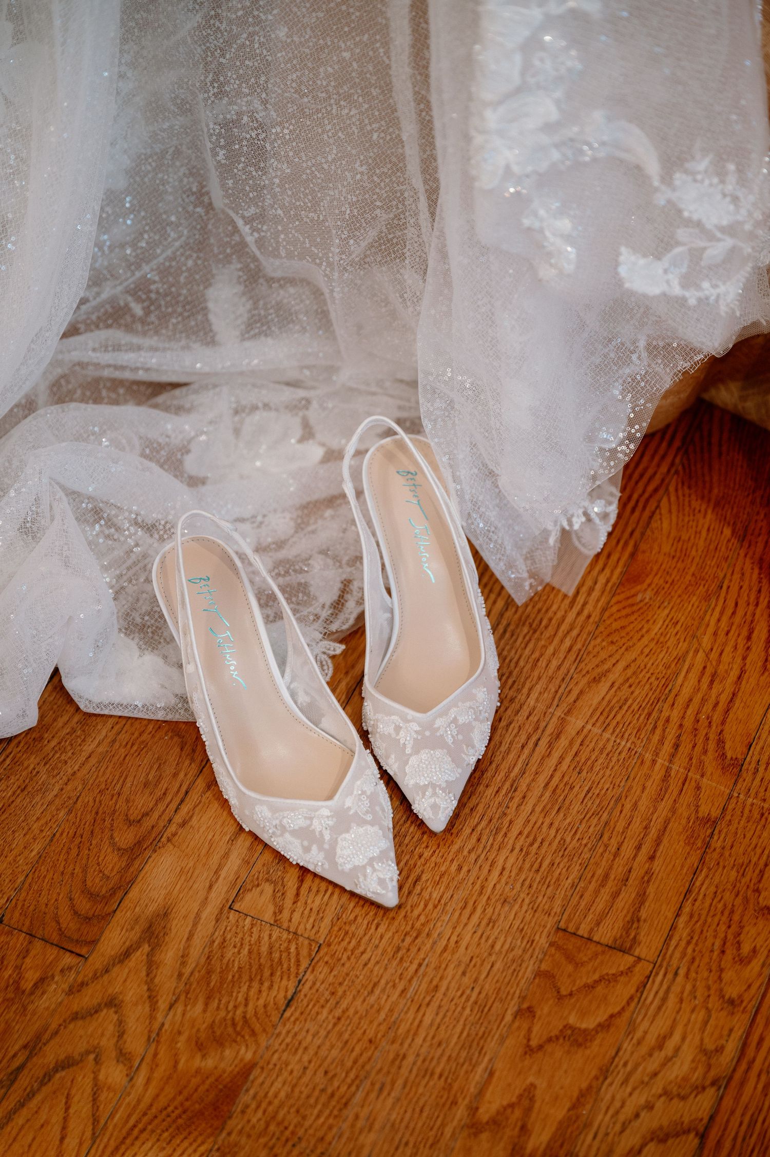 White lace pointed-toe slingback wedding heels resting on hardwood floor beside an elegant lace bridal gown.