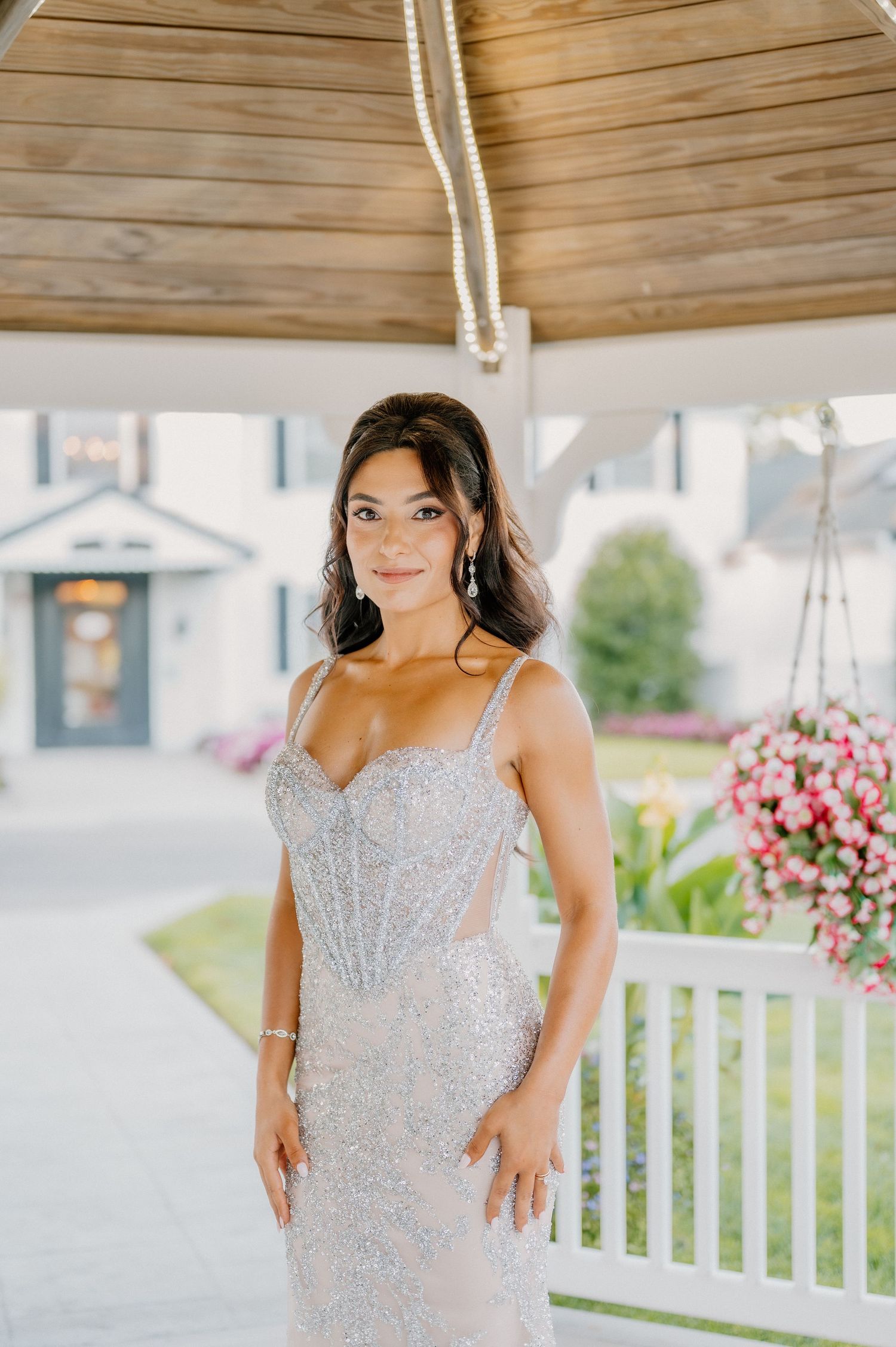 A person in a sparkly silver formal dress poses on a white porch with pink flowers in the background.