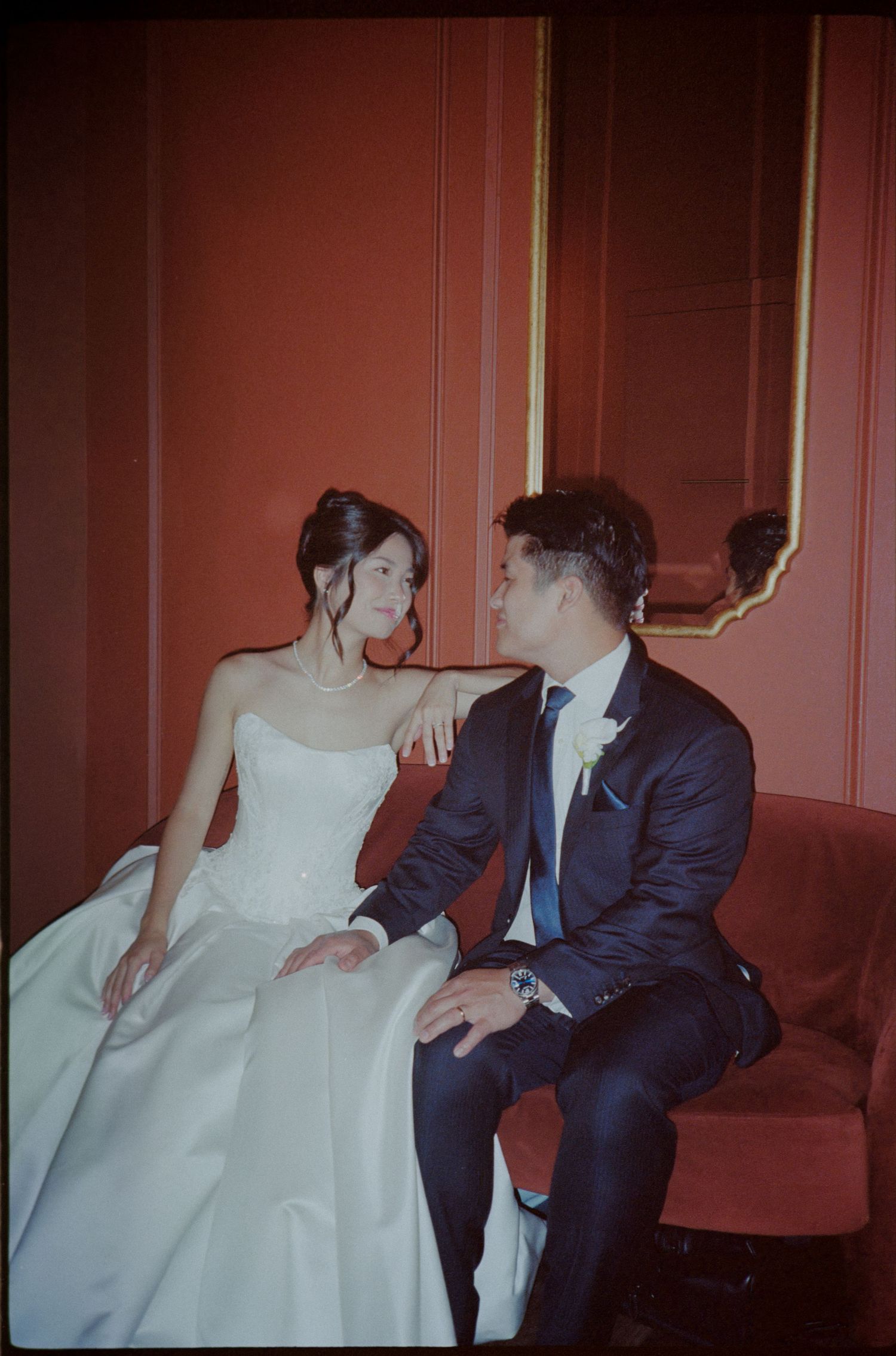 A bride in a white gown and groom in a navy suit sit together on a red velvet sofa in an elegant room.