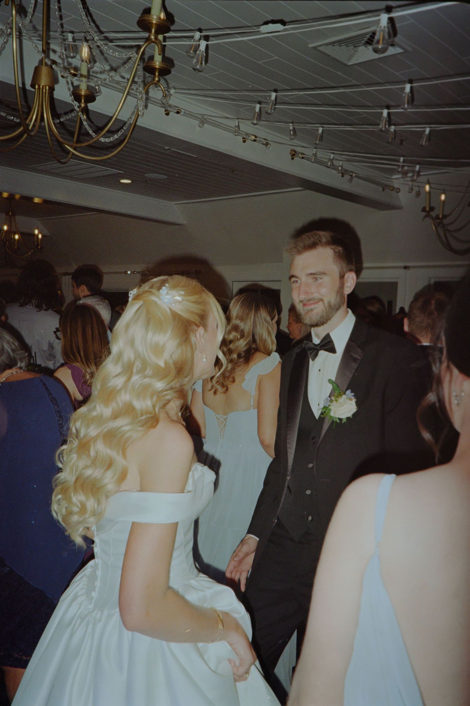 A bride in an off-shoulder white gown dances with a groom in a black tuxedo at a wedding reception.