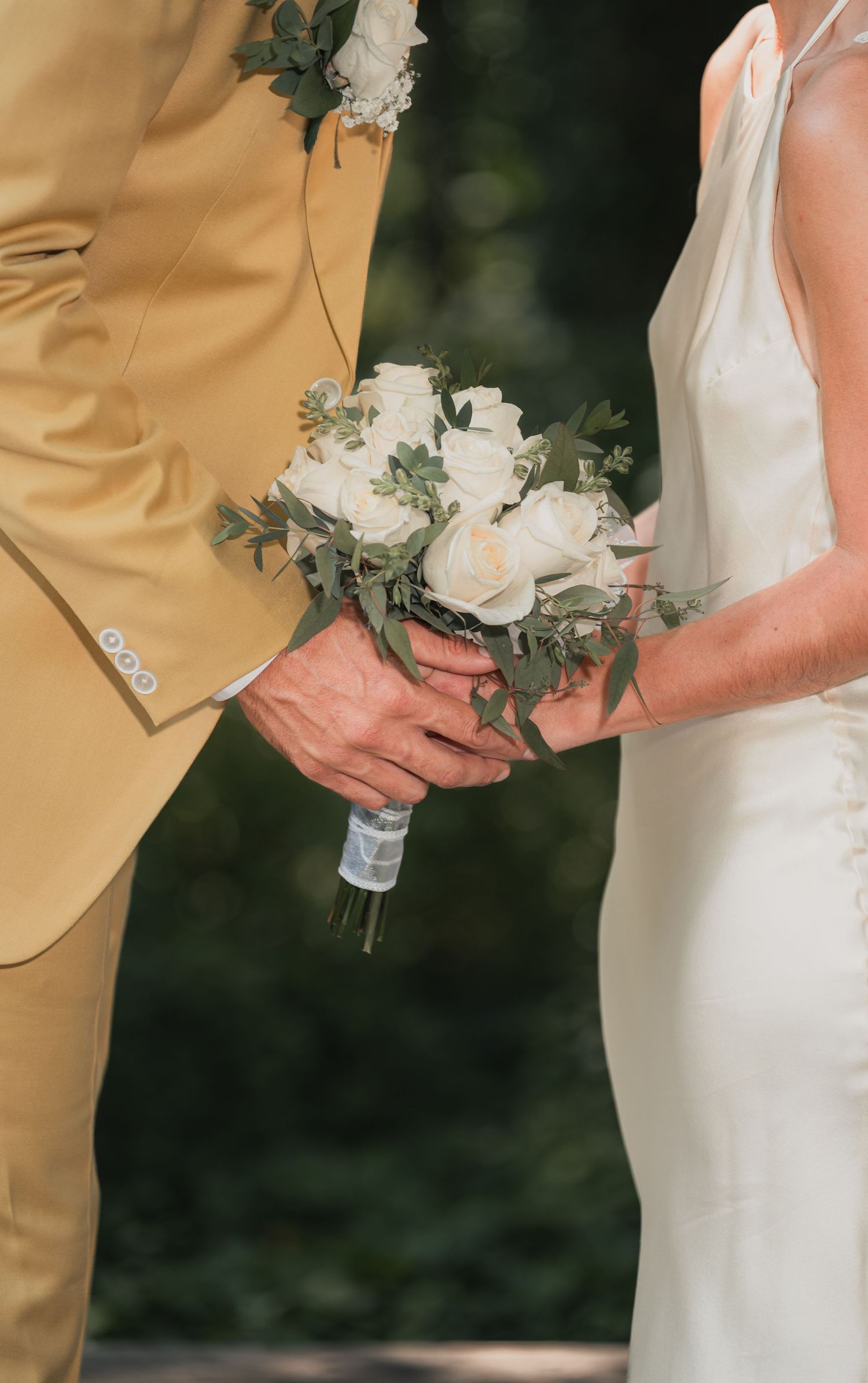 Close-up detail of wedding couple's hands holding white rose bouquets against tan and black formal attire.