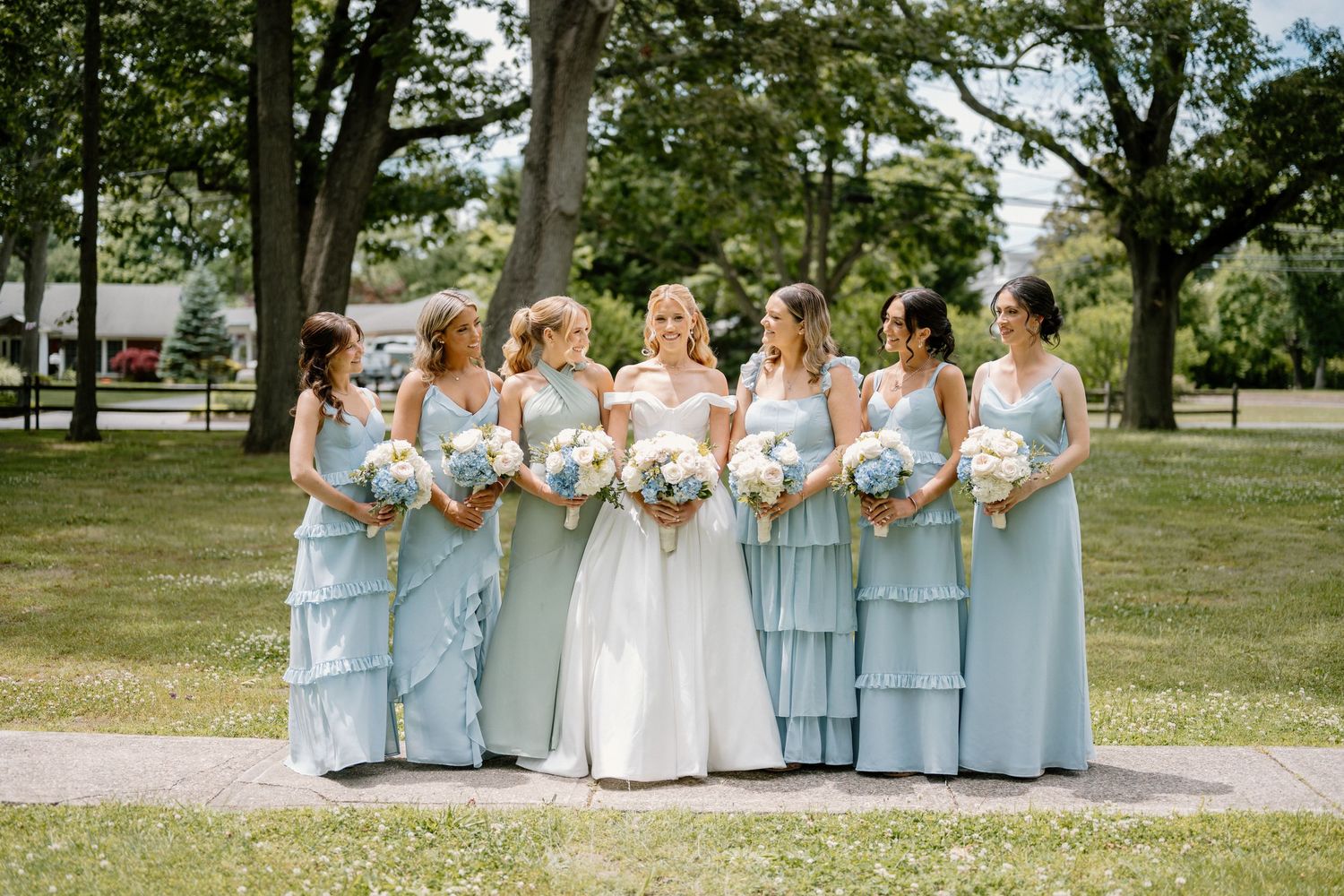 Bride in white gown poses with six bridesmaids in light blue dresses holding white bouquets outdoors on sunny day.