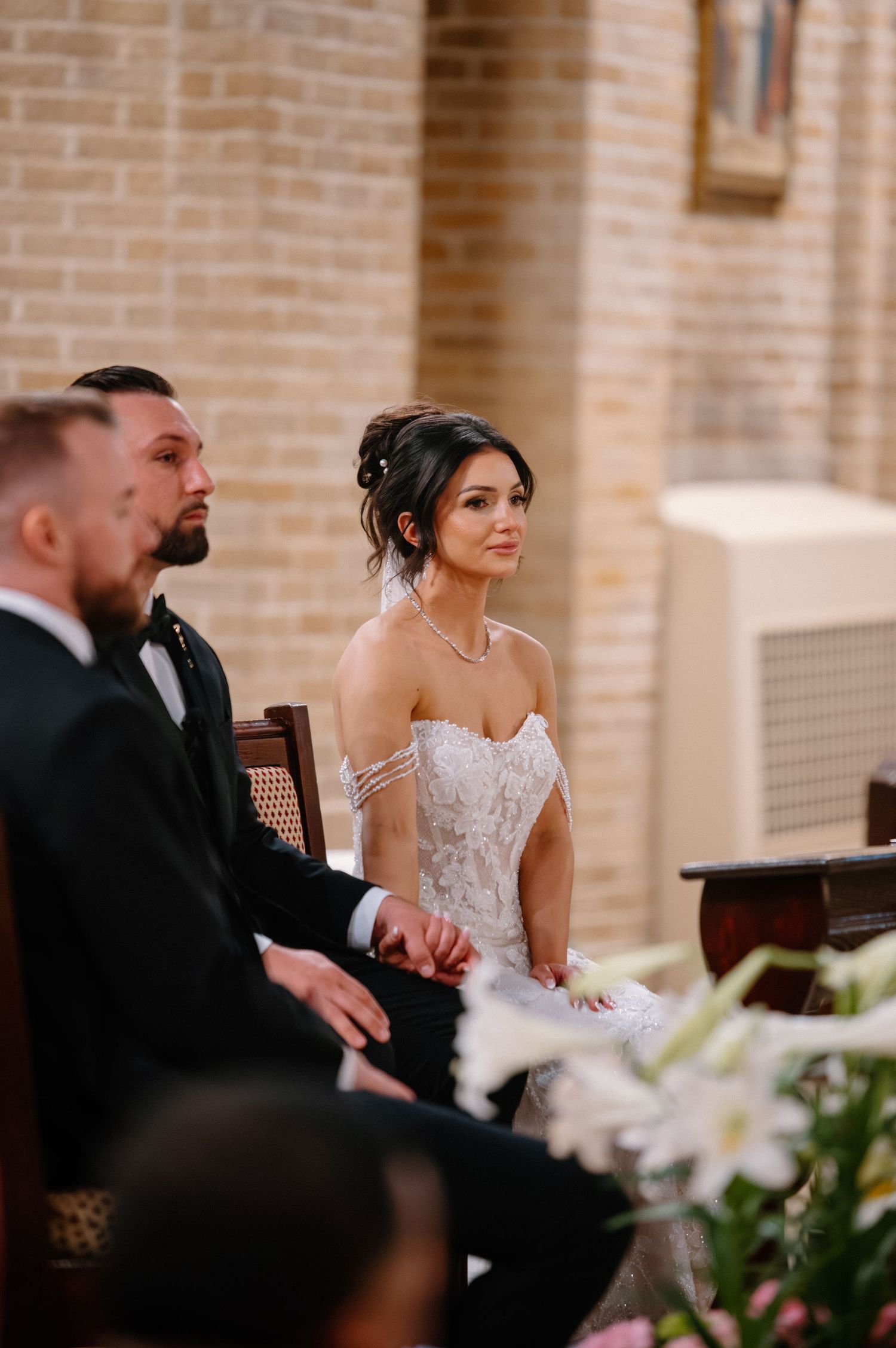 Bride in lace gown and groom in tuxedo seated during wedding ceremony in a brick church.