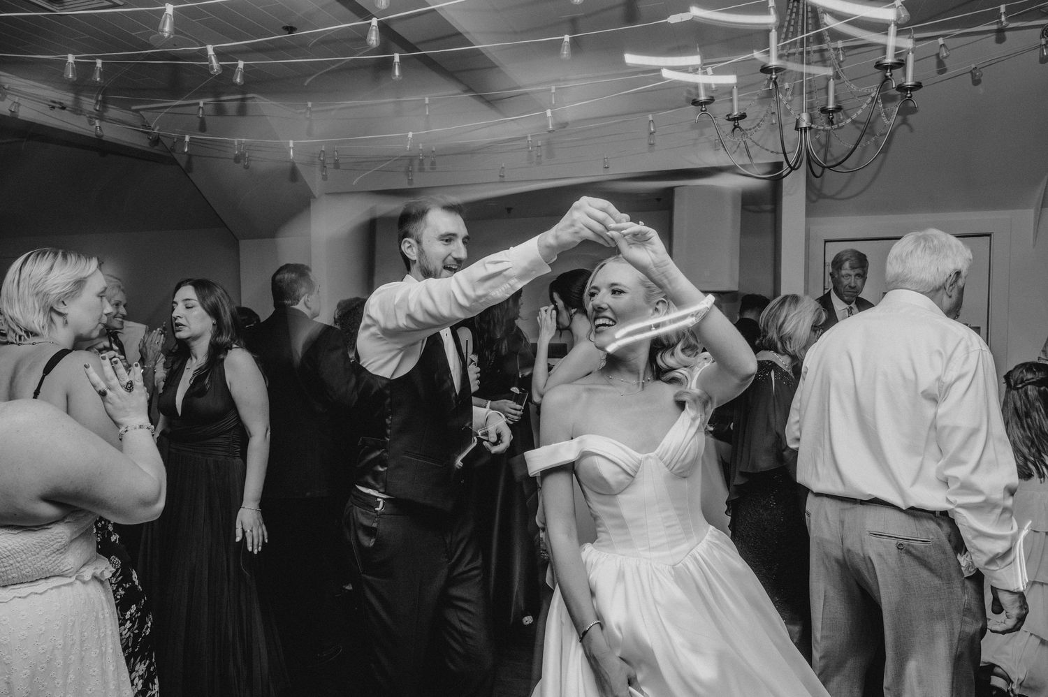 Black and white photo of bride and groom dancing together at wedding reception with guests celebrating in background.