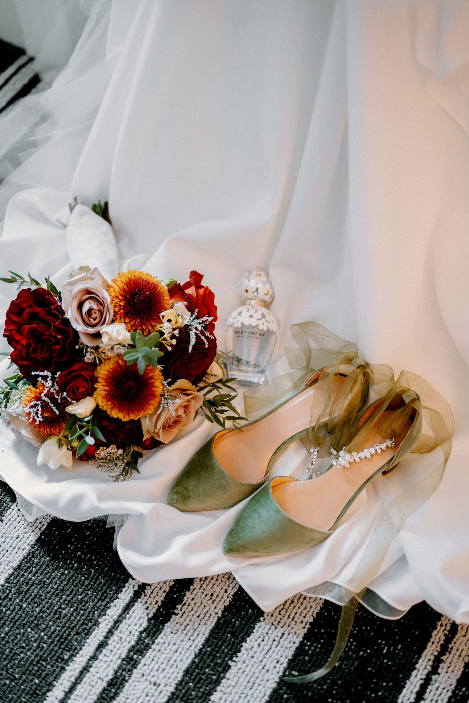 Bridal flat lay with green velvet heels, autumn bouquet of sunflowers and roses, and perfume bottle on white satin fabric.