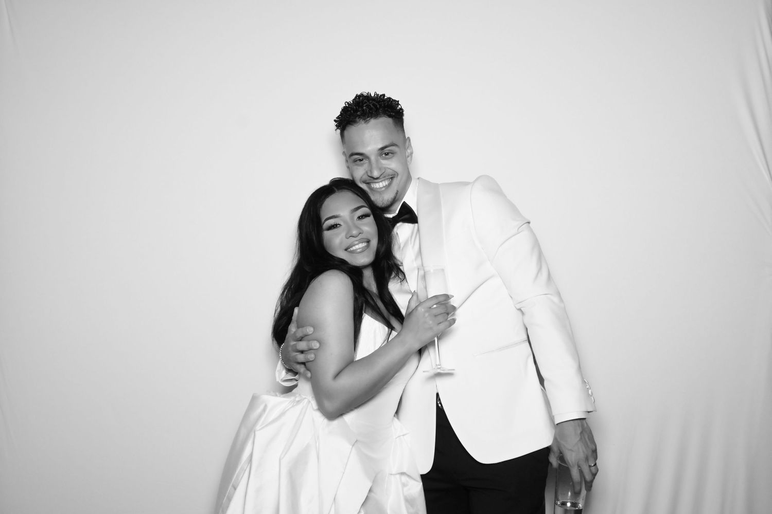 Black and white photo of a smiling couple in formal wedding attire, embracing warmly against a white background.