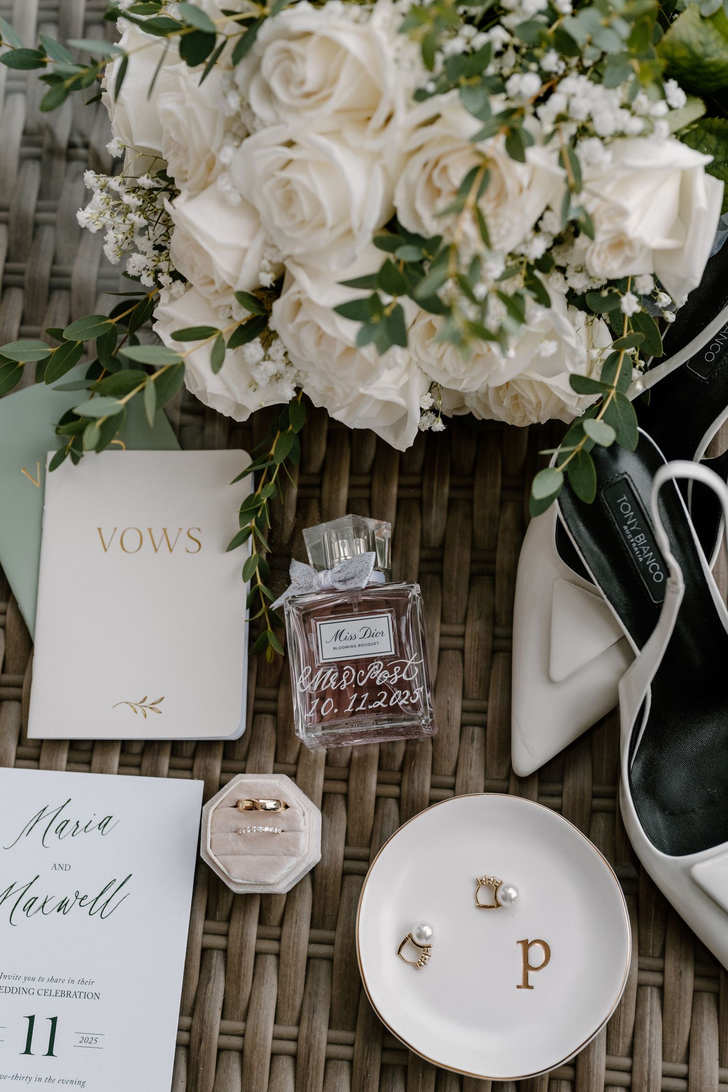 Elegant bridal flat lay with white roses, vow books, Miss Dior perfume, wedding rings, pearl earrings, and white heels.