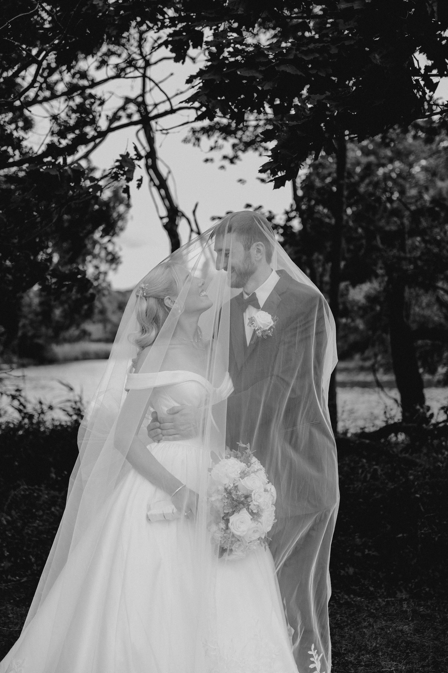 Black and white wedding photo of bride and groom sharing a kiss under a flowing veil outdoors.