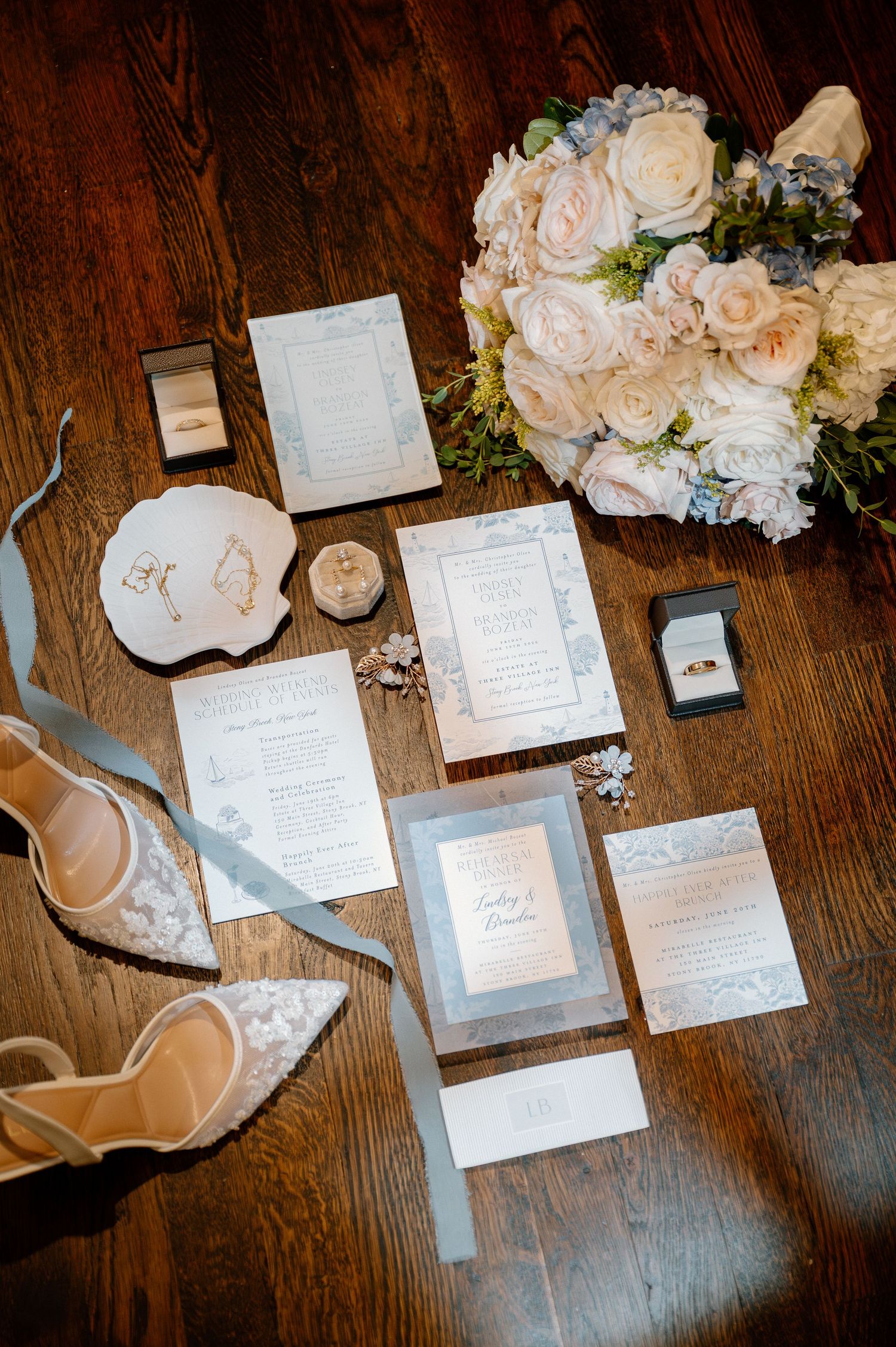 Elegant wedding flat lay with floral invitations, blush bouquet, lace heels, rings, and dusty blue ribbons on wood floor.