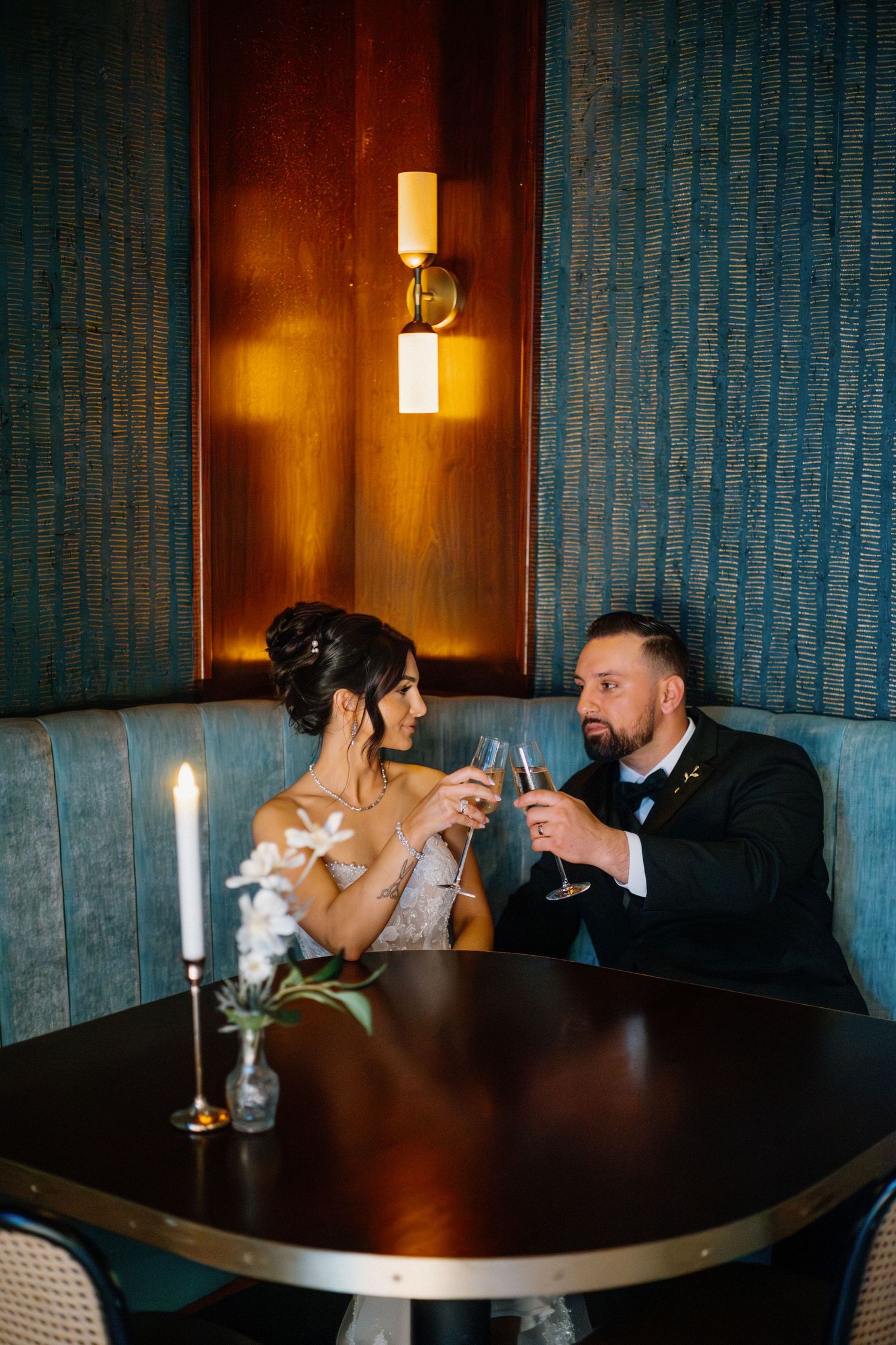 Bride and groom toasting champagne glasses at a candlelit booth, dressed in wedding attire at an elegant venue.