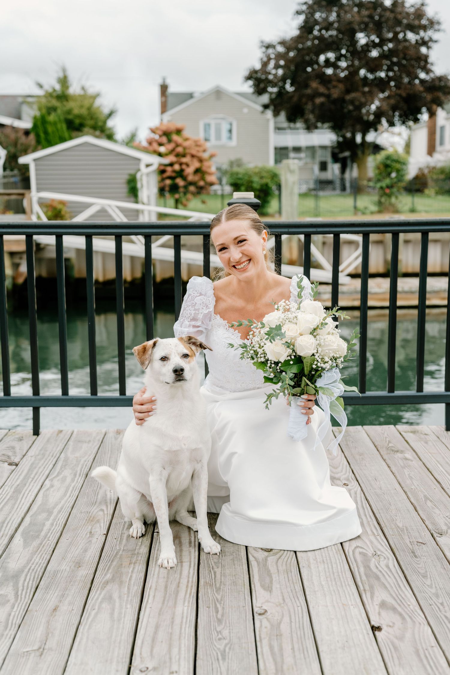 A white wedding dress and bouquet photographed with a small white dog on a wooden deck.