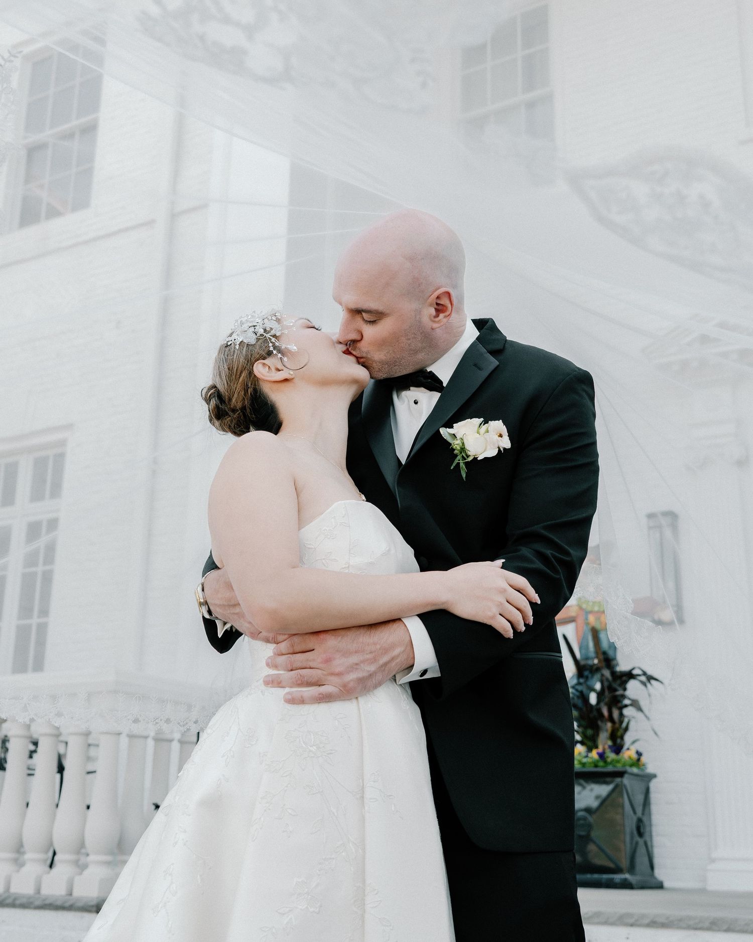 A romantic wedding couple shares tender moments on a balcony with white columns in formal attire.