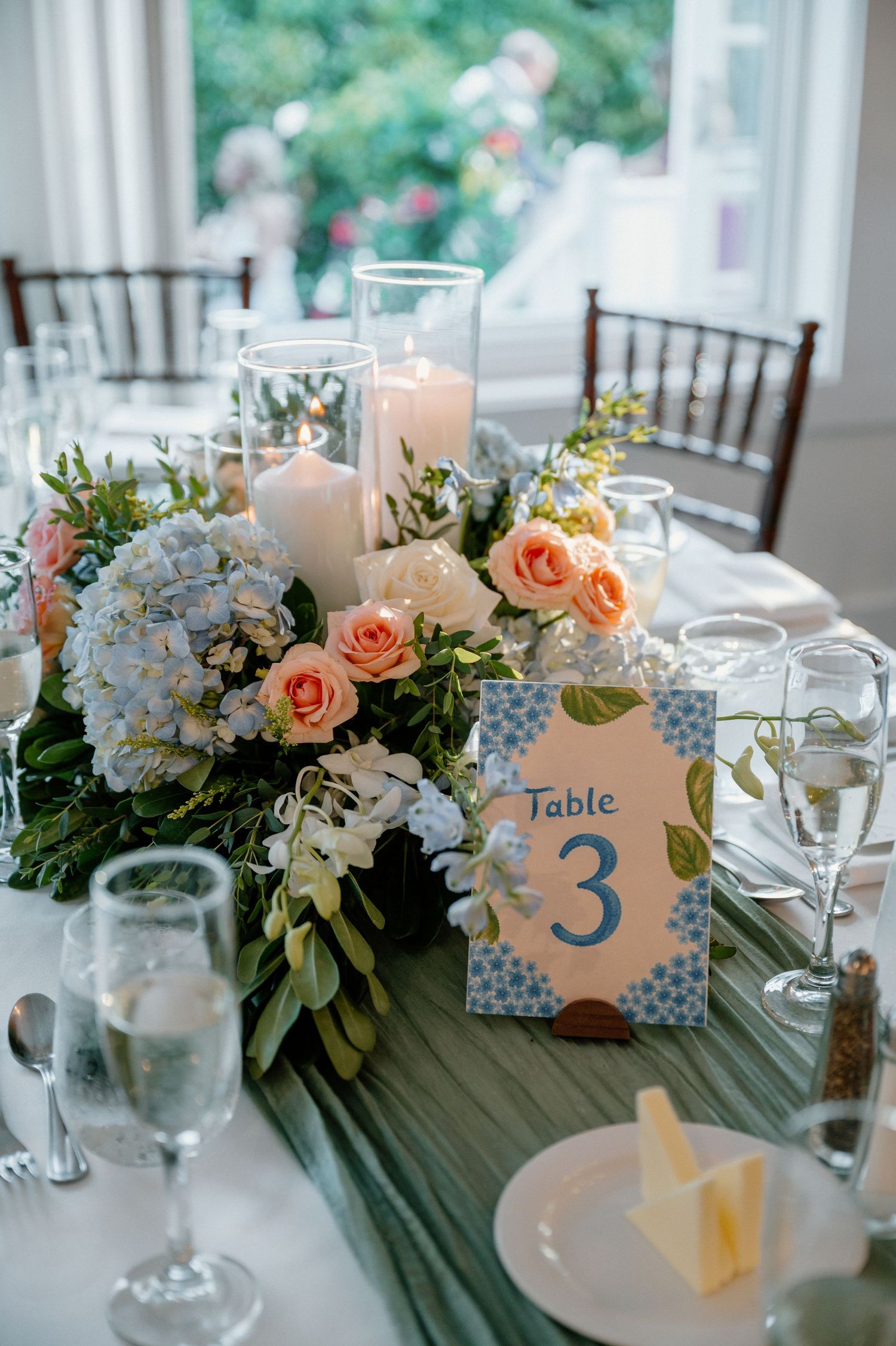Wedding table centerpiece with blue hydrangeas, peach roses, white candles, and a Table 3 card on an elegant white tablecloth.