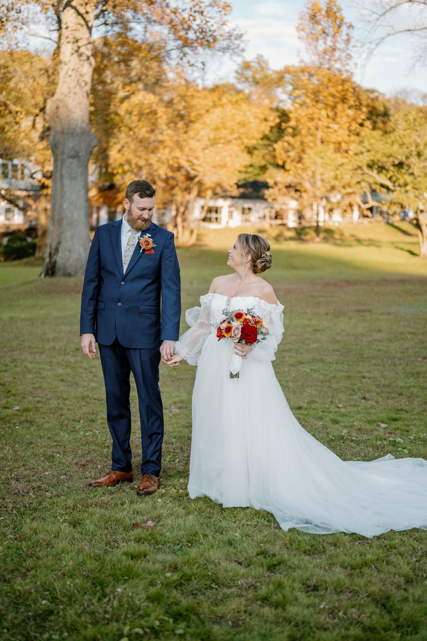 A couple in wedding attire stand together on a grassy lawn surrounded by autumn trees with golden foliage.