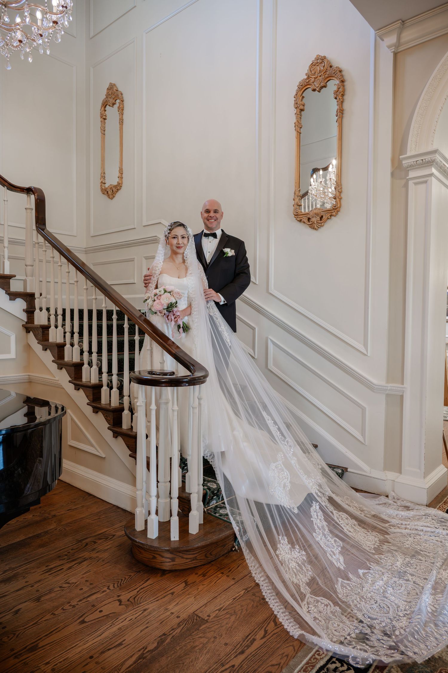 An elegant wedding photo on a grand staircase with ornate gold mirrors and a stunning cathedral-length lace veil.
