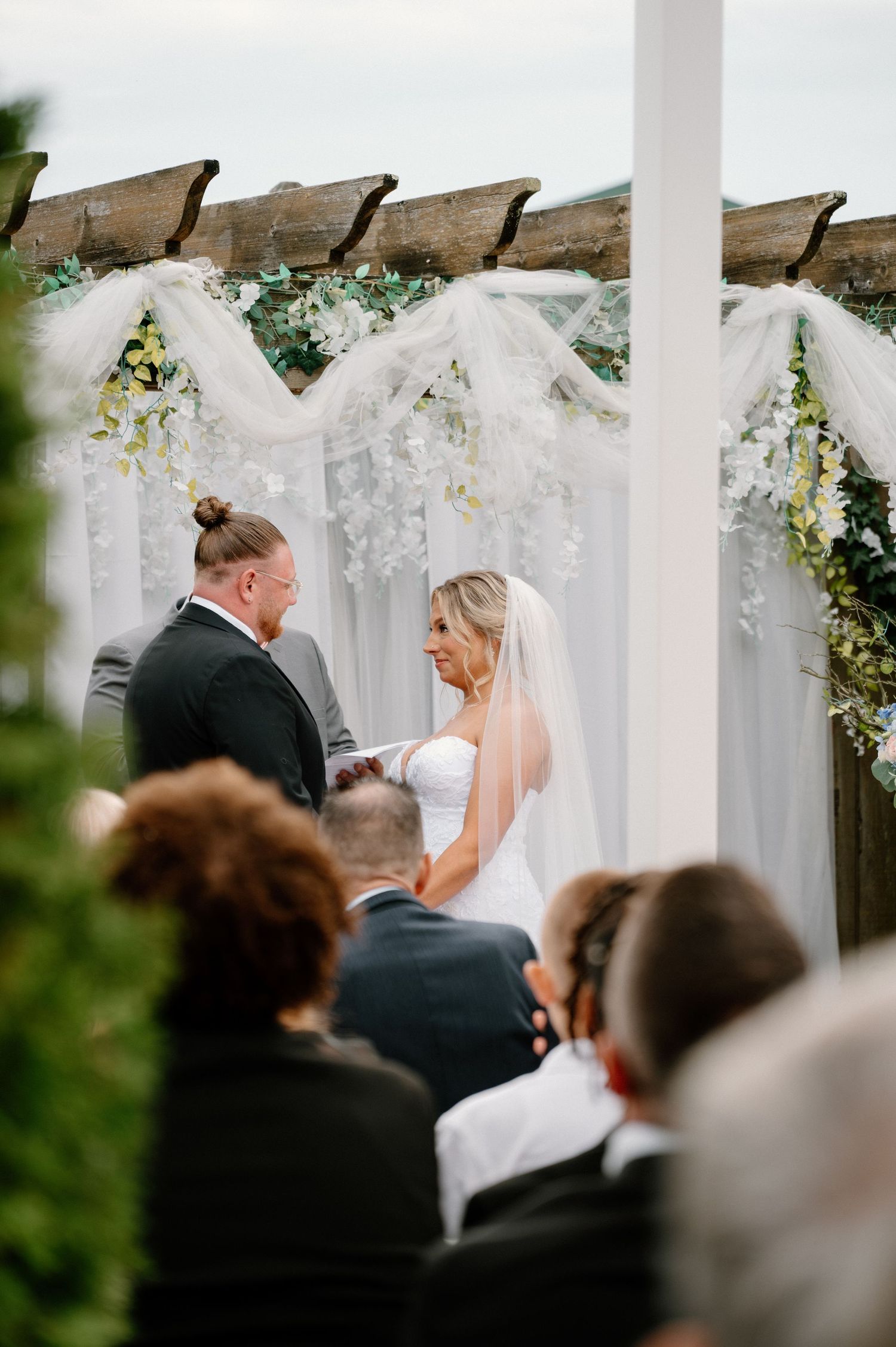Outdoor wedding ceremony under a white pergola decorated with floral arrangements.