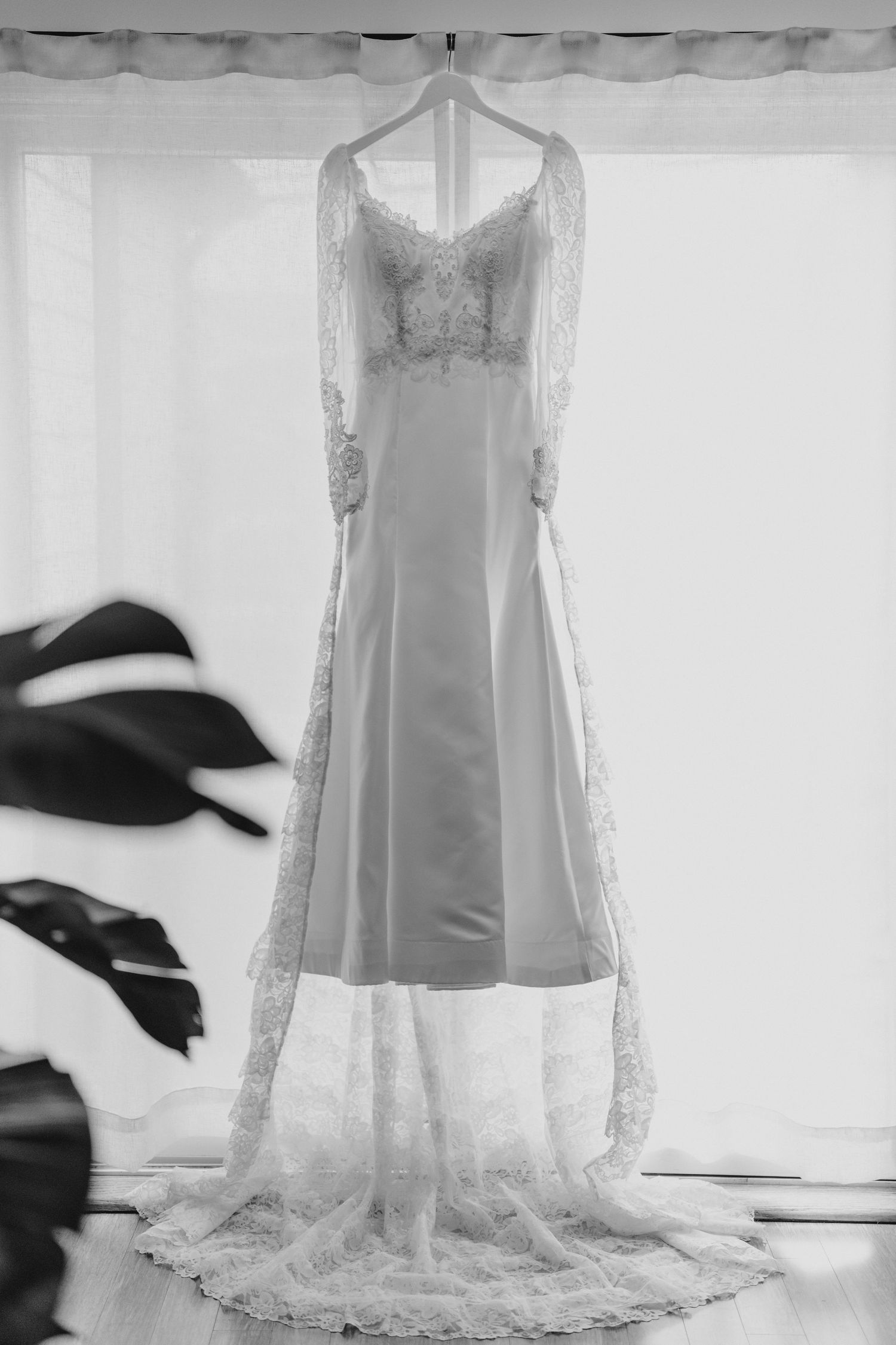 Elegant wedding dress with lace sleeves and train hanging on a hanger in front of a window, black and white photo.