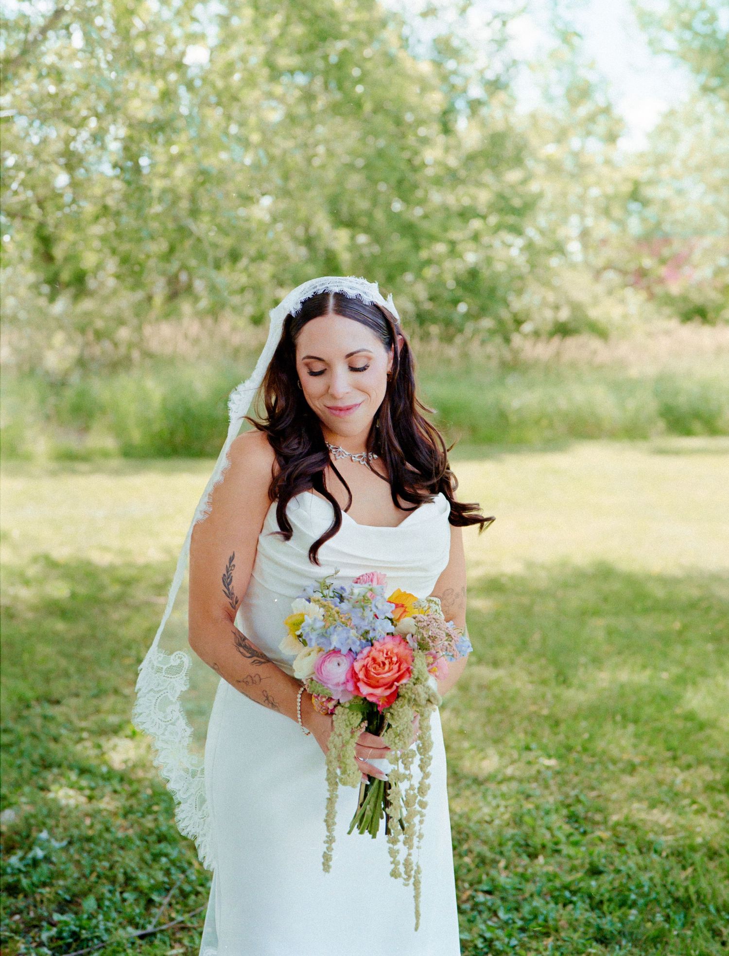 Bride in white gown and lace veil holds colorful wildflower bouquet outdoors on sunny day.