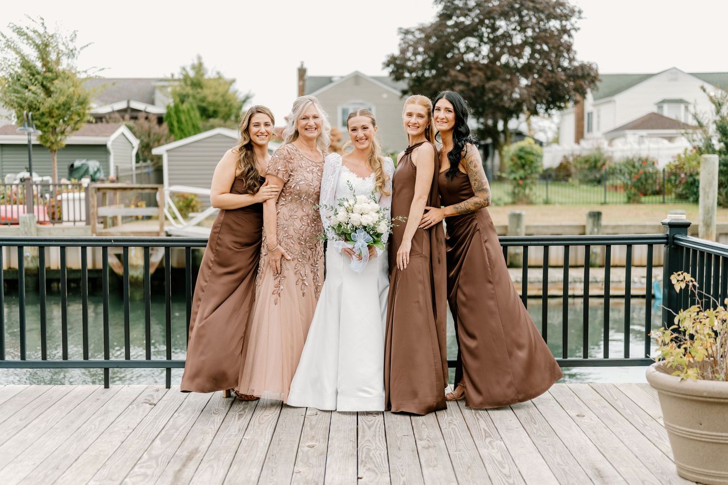Wedding party members in brown and blush dresses pose together on a deck overlooking water.