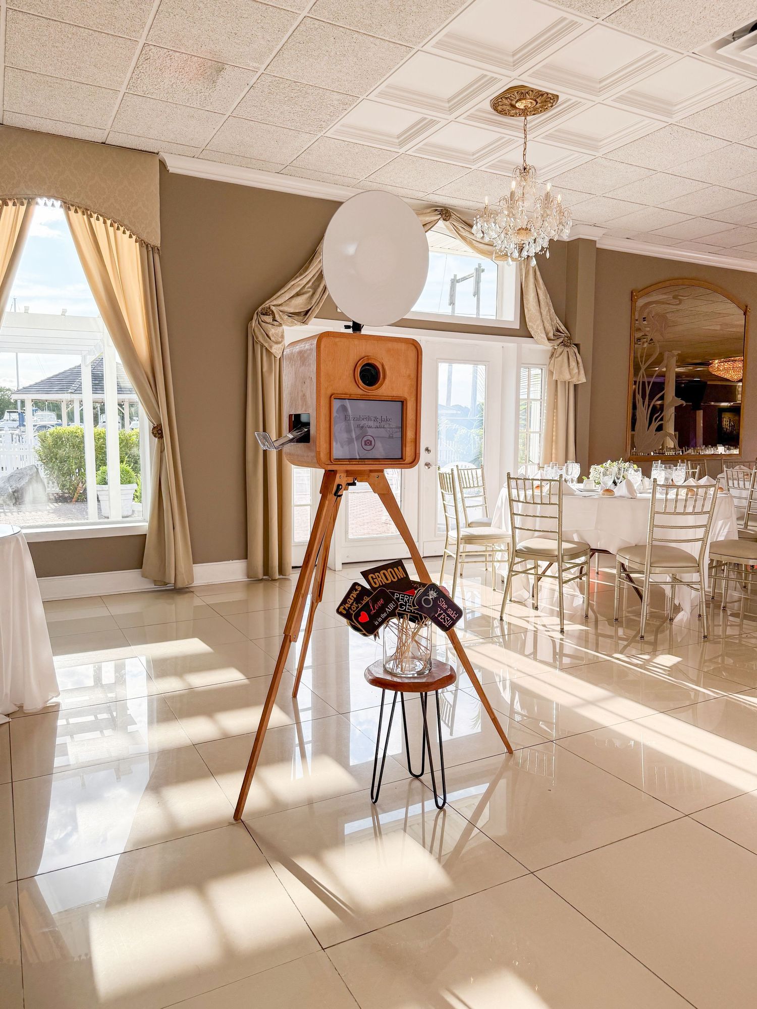Wooden photo booth setup with circular light in an elegant banquet hall with chandelier and drapes.