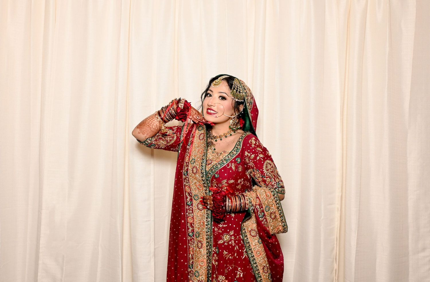 Traditional South Asian bride in ornate red and gold wedding attire poses against light backdrop.